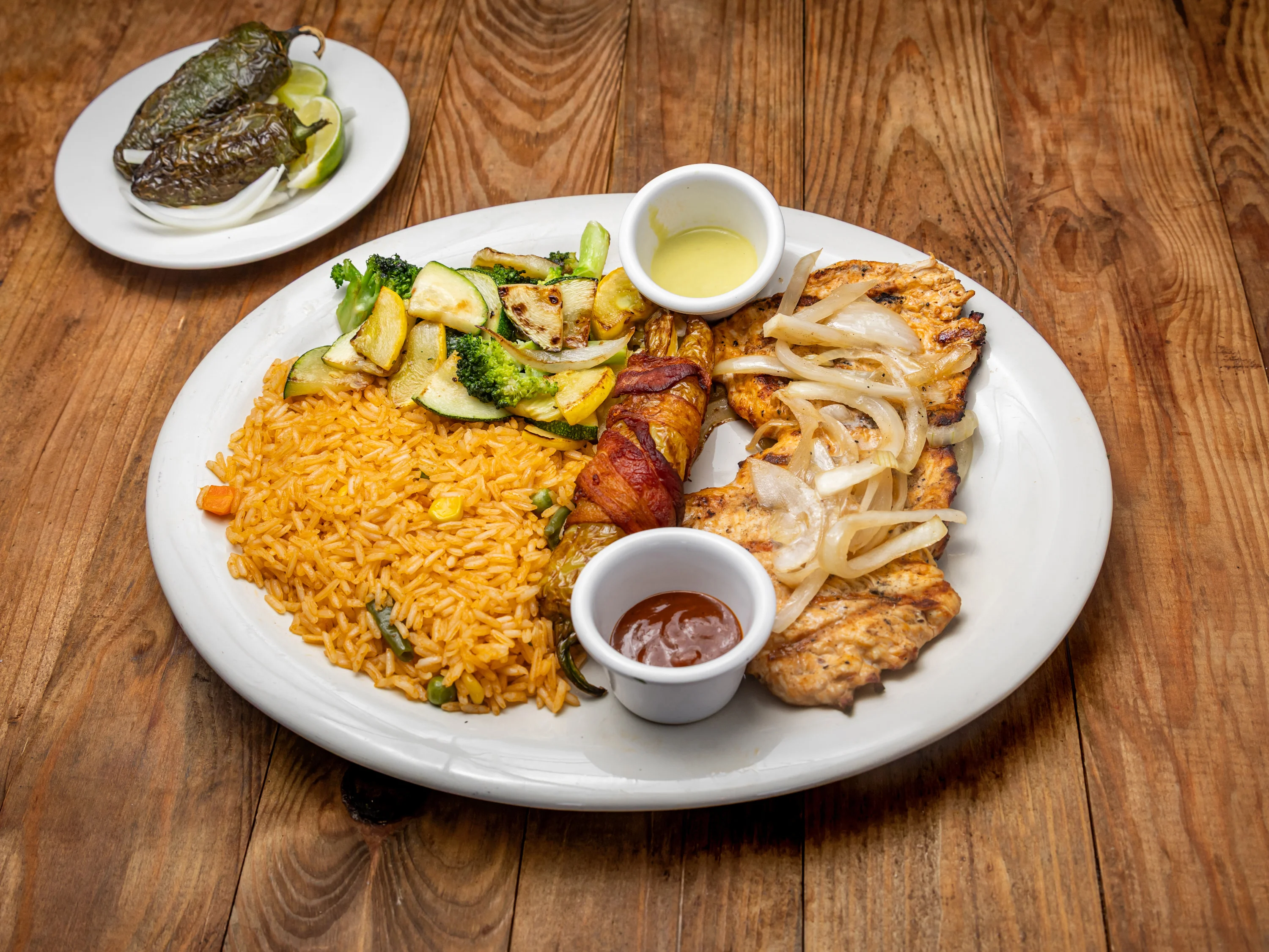 A plate of grilled chicken with rice, grilled vegetables, and dipping sauces, accompanied by jalapeños and lime wedges.