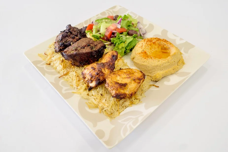 A hearty meal featuring grilled meats, rice, a fresh salad with tomatoes and greens, and a side of hummus, all served on a patterned plate.