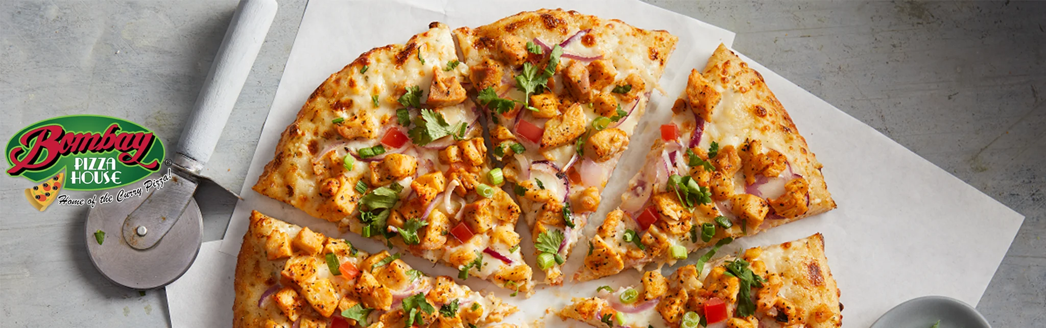 A close-up of a pizza topped with chicken, red onions, tomatoes, and herbs, cut into slices, with a pizza cutter beside it.