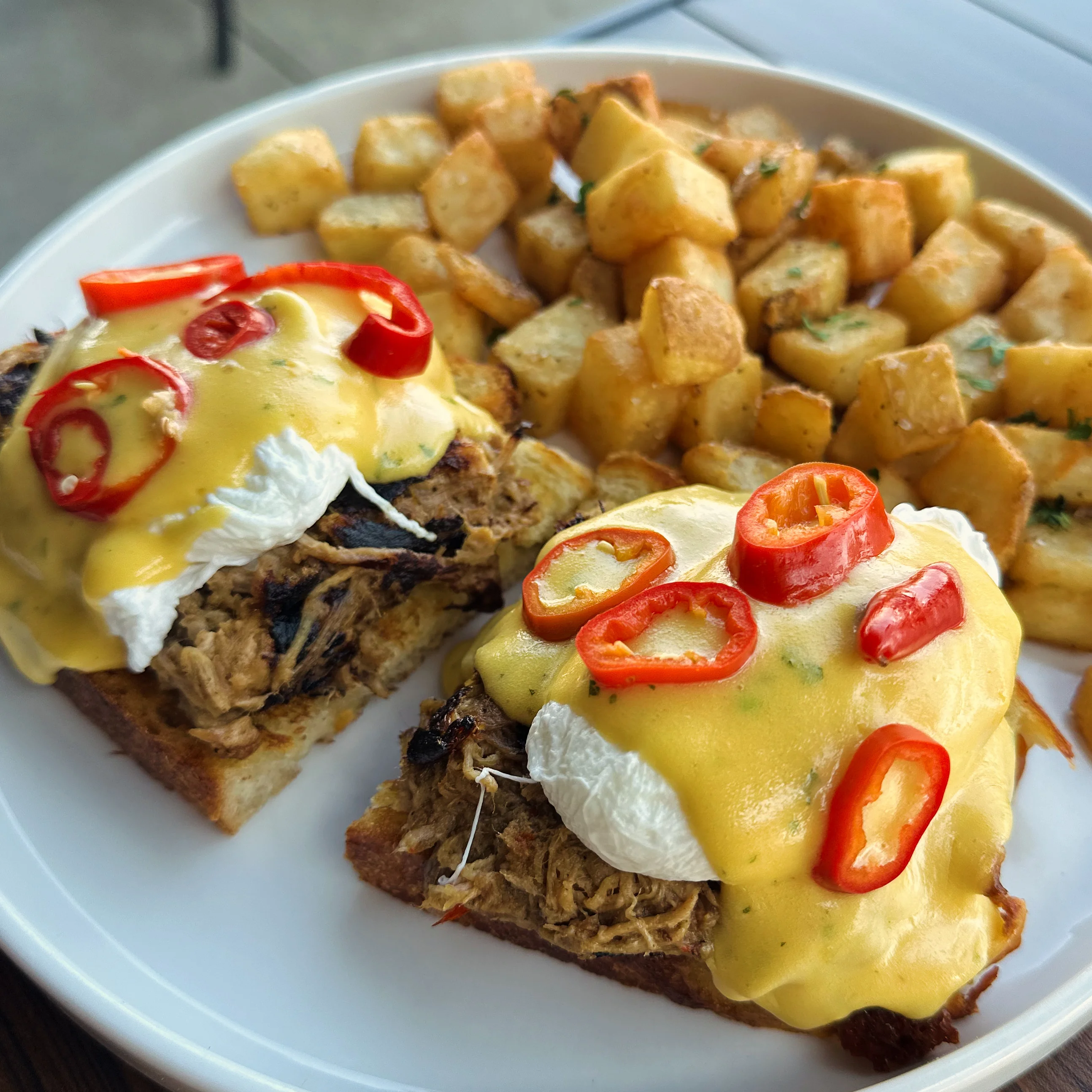 A delicious plate of pulled pork eggs benedict served with crispy potato cubes and topped with red chili slices.
