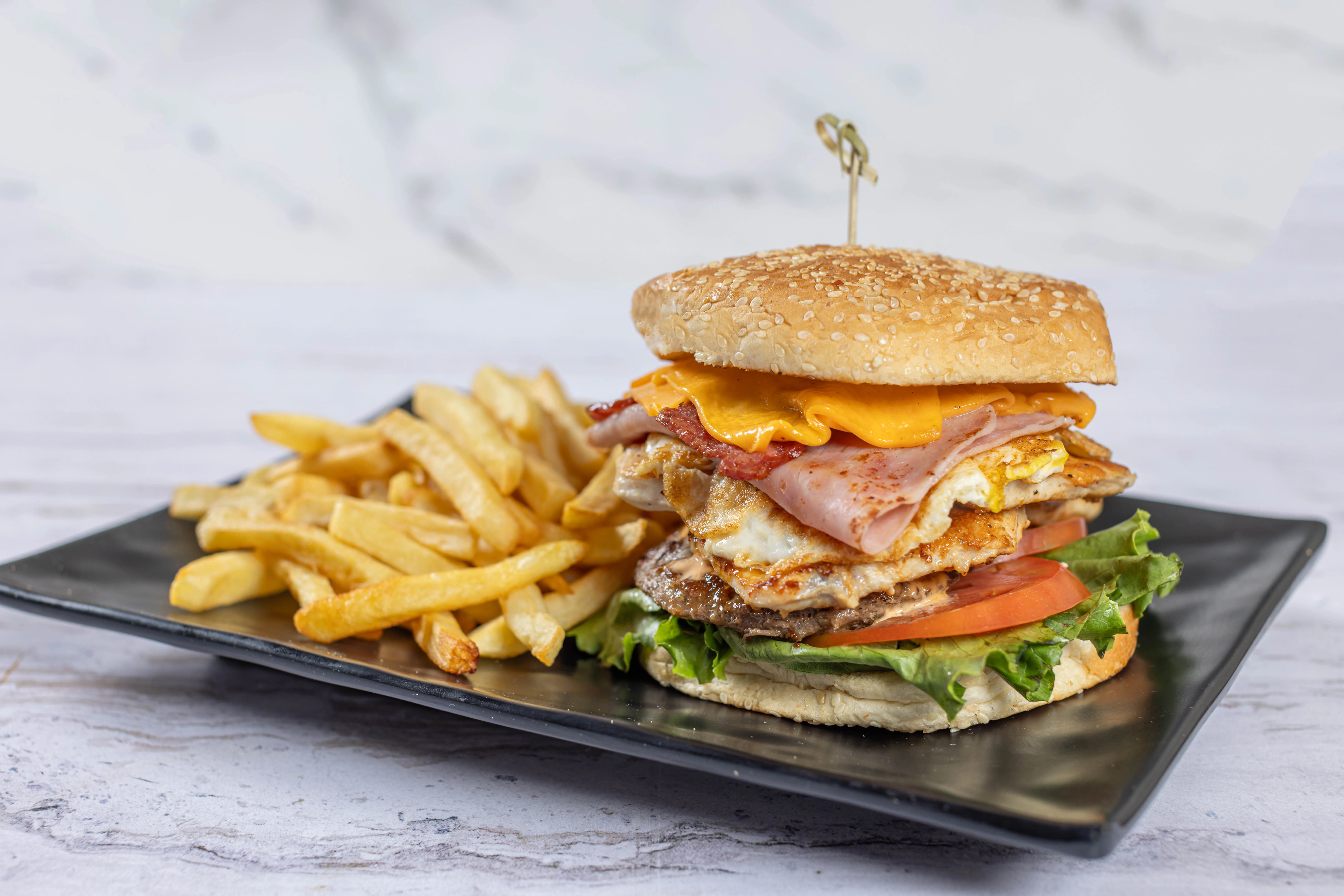 A fully loaded Latin-style hamburger from El Budare Cafe with ham, bacon, and a fried egg, served with a side of french fries.