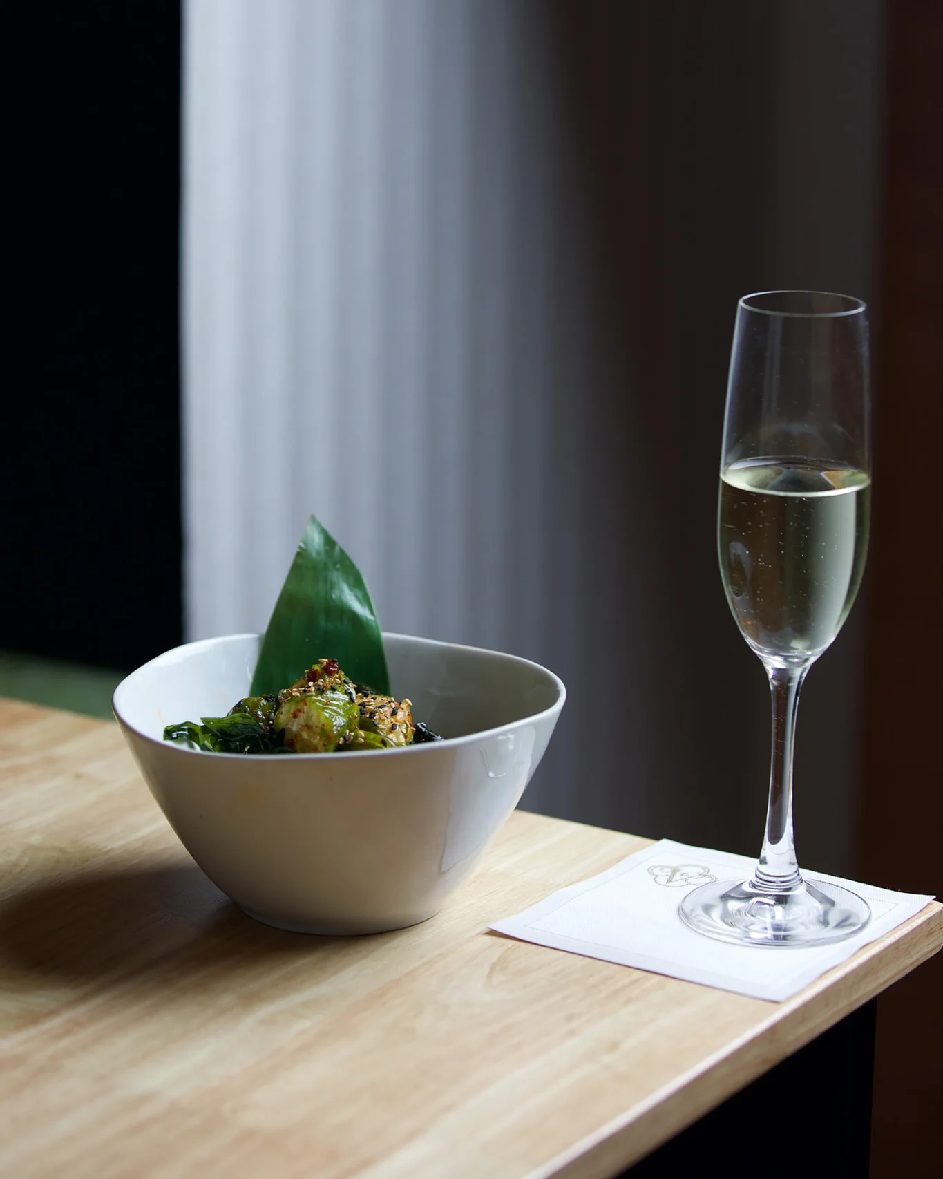 A white ceramic bowl containing roasted Brussels sprouts garnished with sesame seeds and a green leaf, sitting on a light wooden table next to a filled champagne flute on a white square napkin.