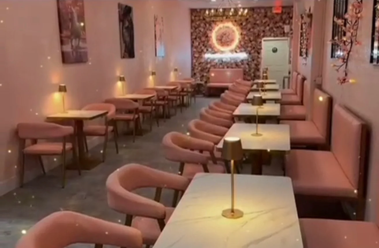 An elegant dining area featuring pastel pink furniture, marble-topped tables, and subtle lighting. The walls are adorned with floral decor, creating a cozy ambiance.
