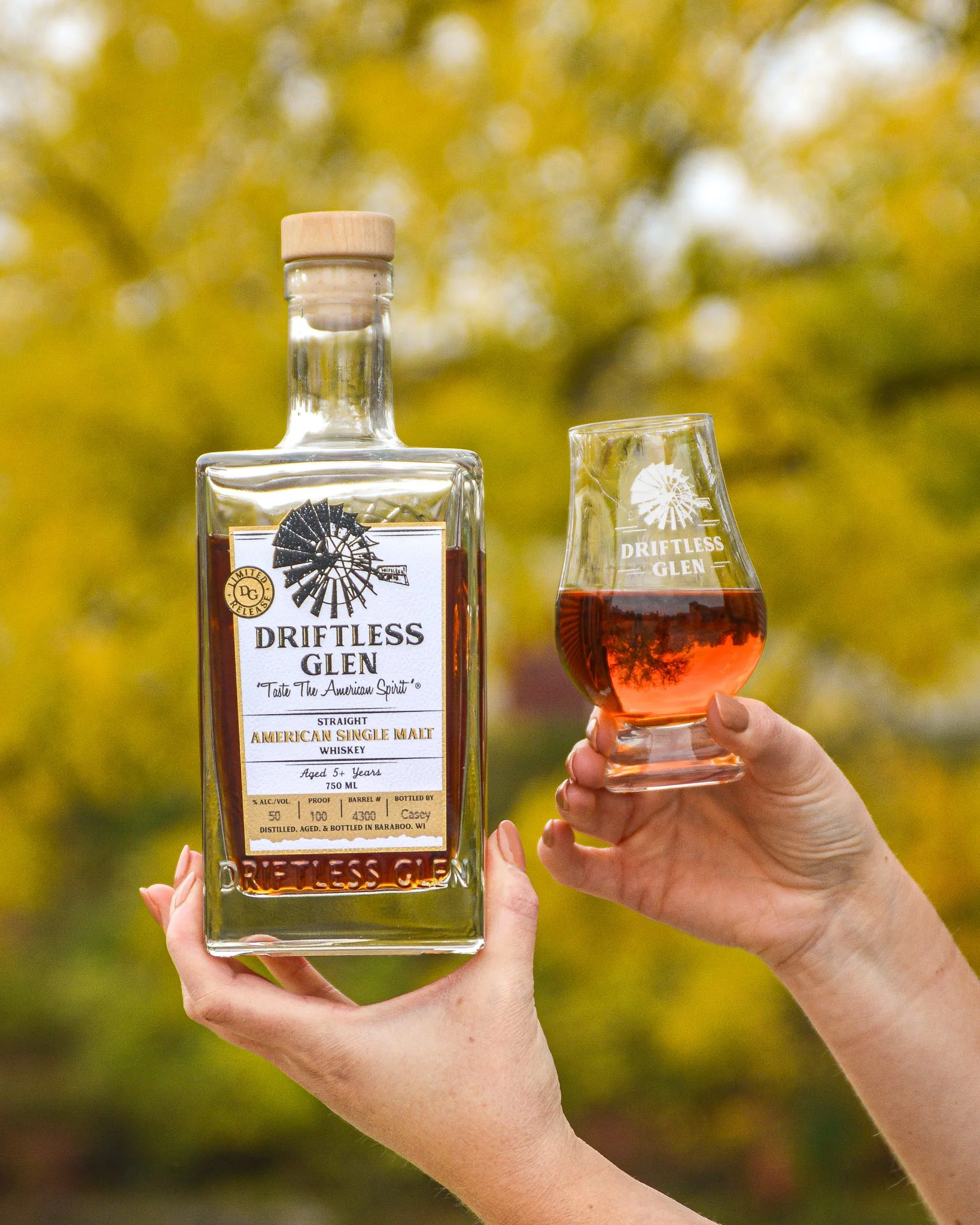 Someone holds up a bottle of American Single Malt and a glass of it poured into, both in front of green trees