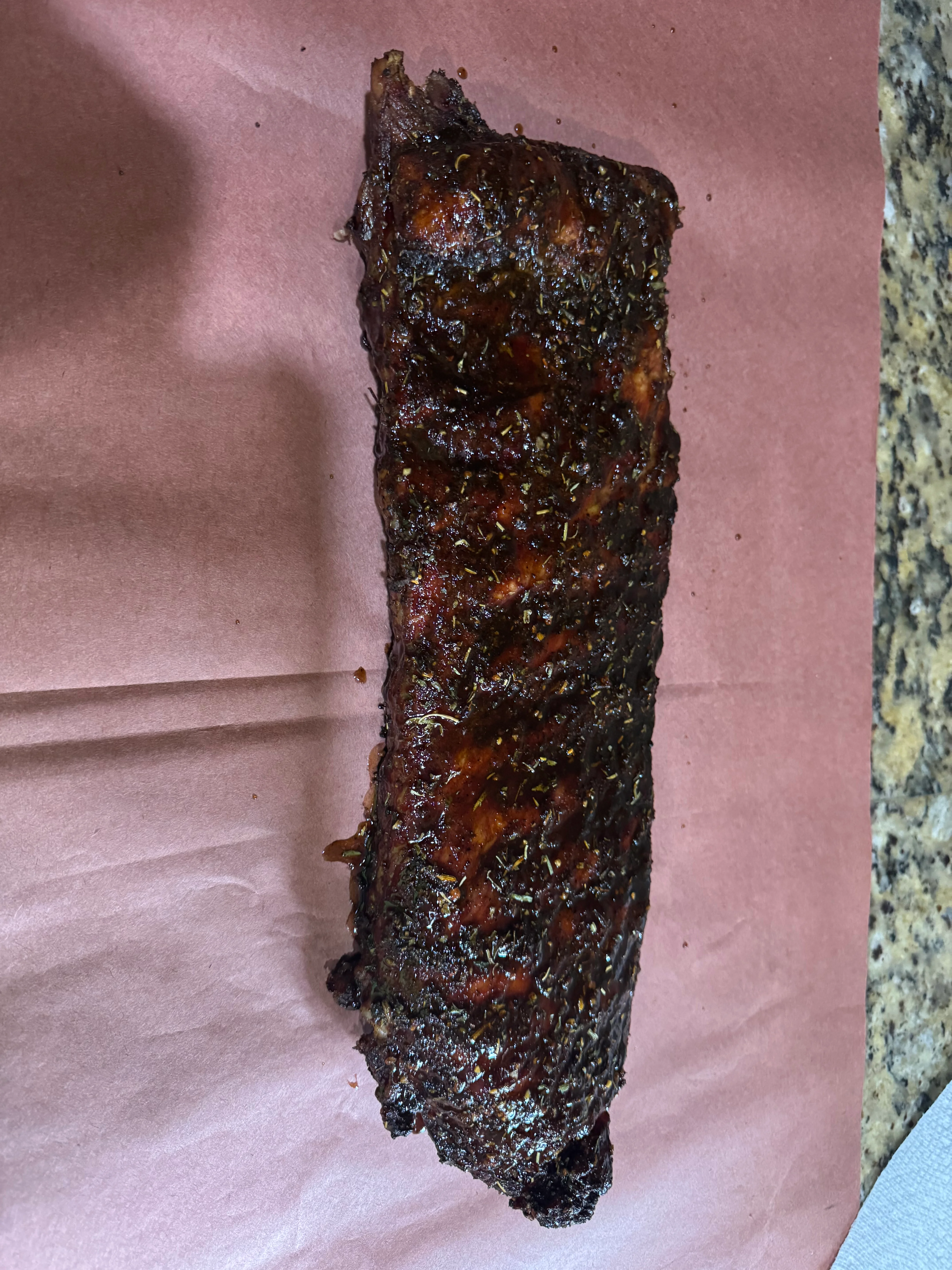 A single rack of glazed barbecue ribs, showcasing a dark, caramelized exterior sprinkled with herbs, resting on butcher paper.