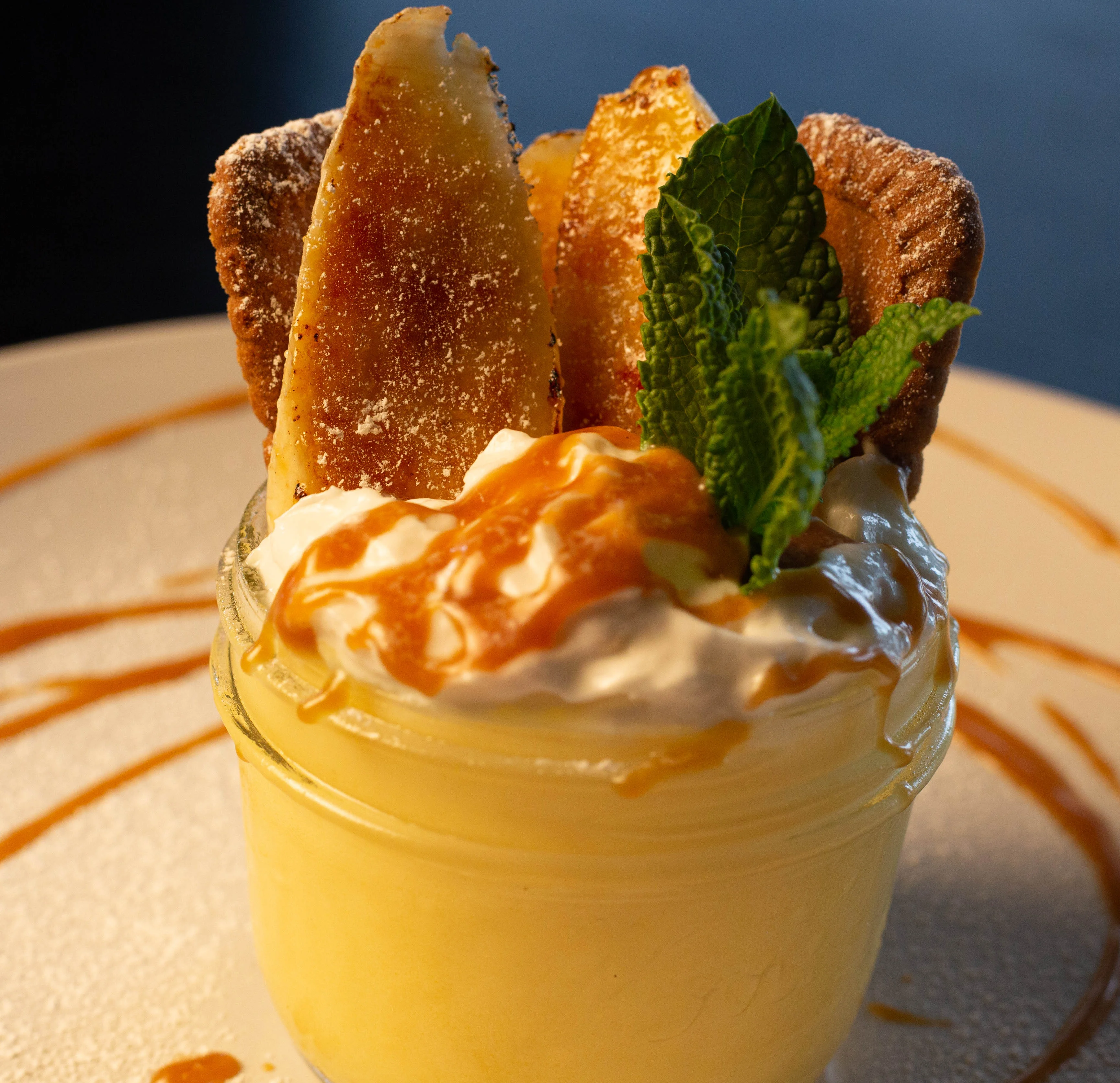 A dessert served in a jar featuring a creamy filling topped with caramel sauce, whipped cream, fried banana slices, and mint leaves.