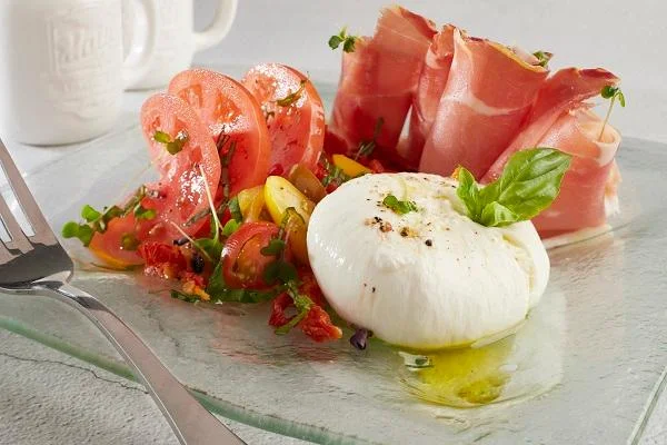 A glass plate featuring fresh tomatoes, prosciutto, and a ball of burrata cheese drizzled with olive oil and garnished with herbs.