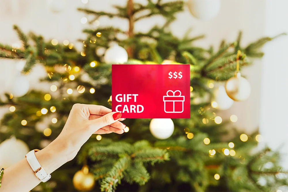 Hand holding a gift card in front of a decorated Christmas tree