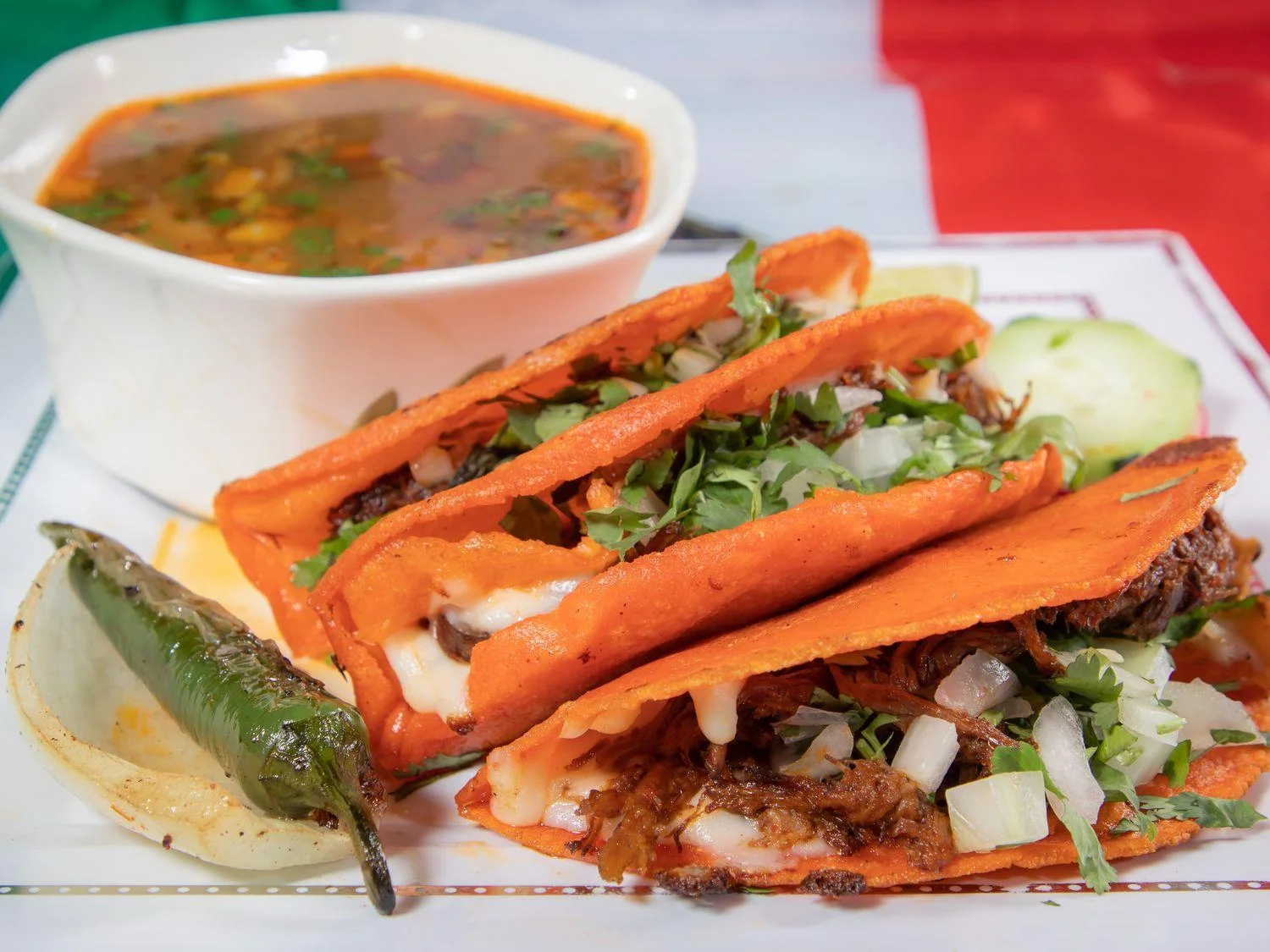 A plate of colorful tacos filled with meat, onions, and cilantro, served with a side of soup and a green chili.