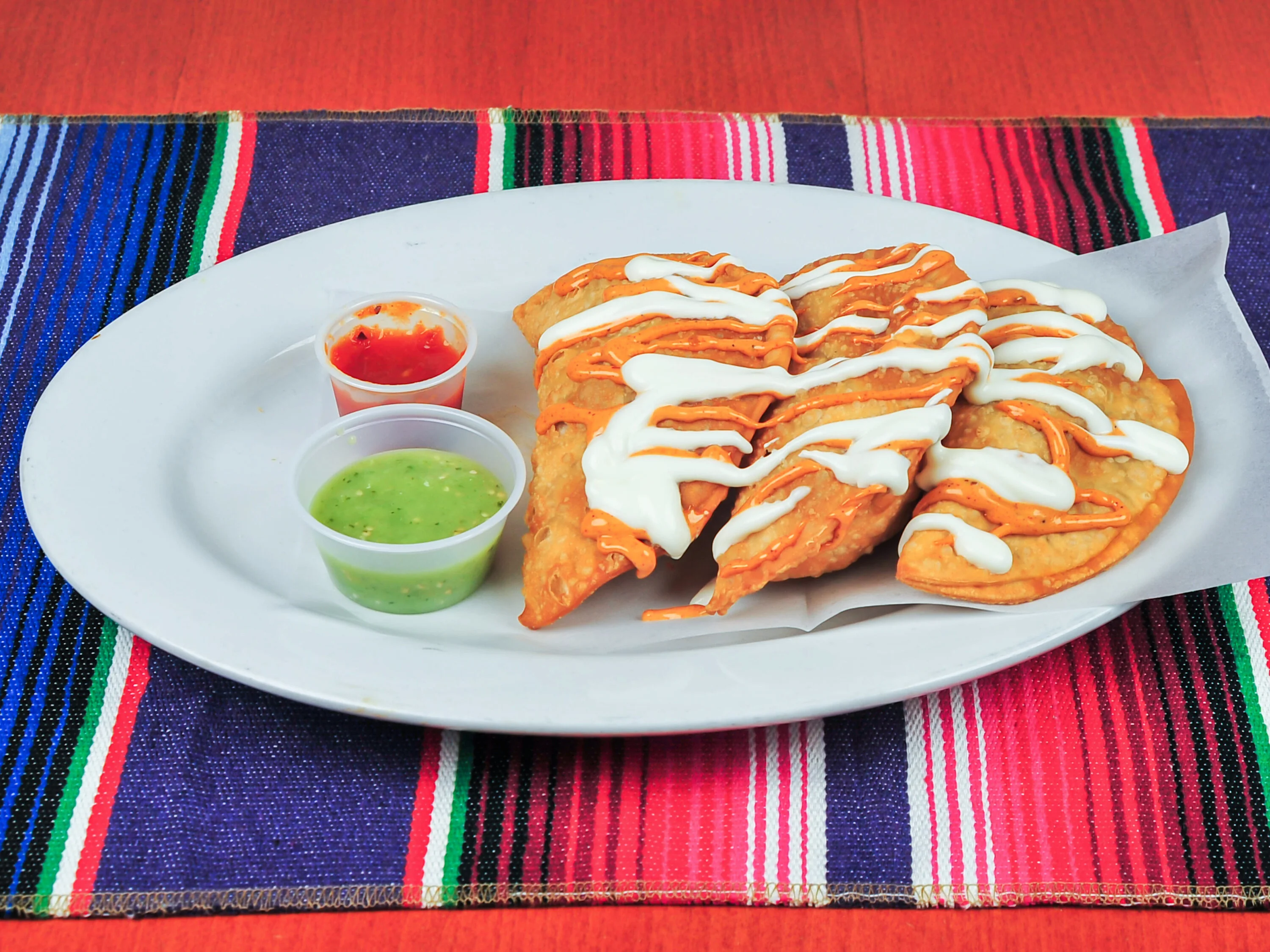 A platter of crispy empanadas drizzled with white sauce and accompanied by two dipping sauces, one red and one green. The plate is set on a colorful woven table runner.