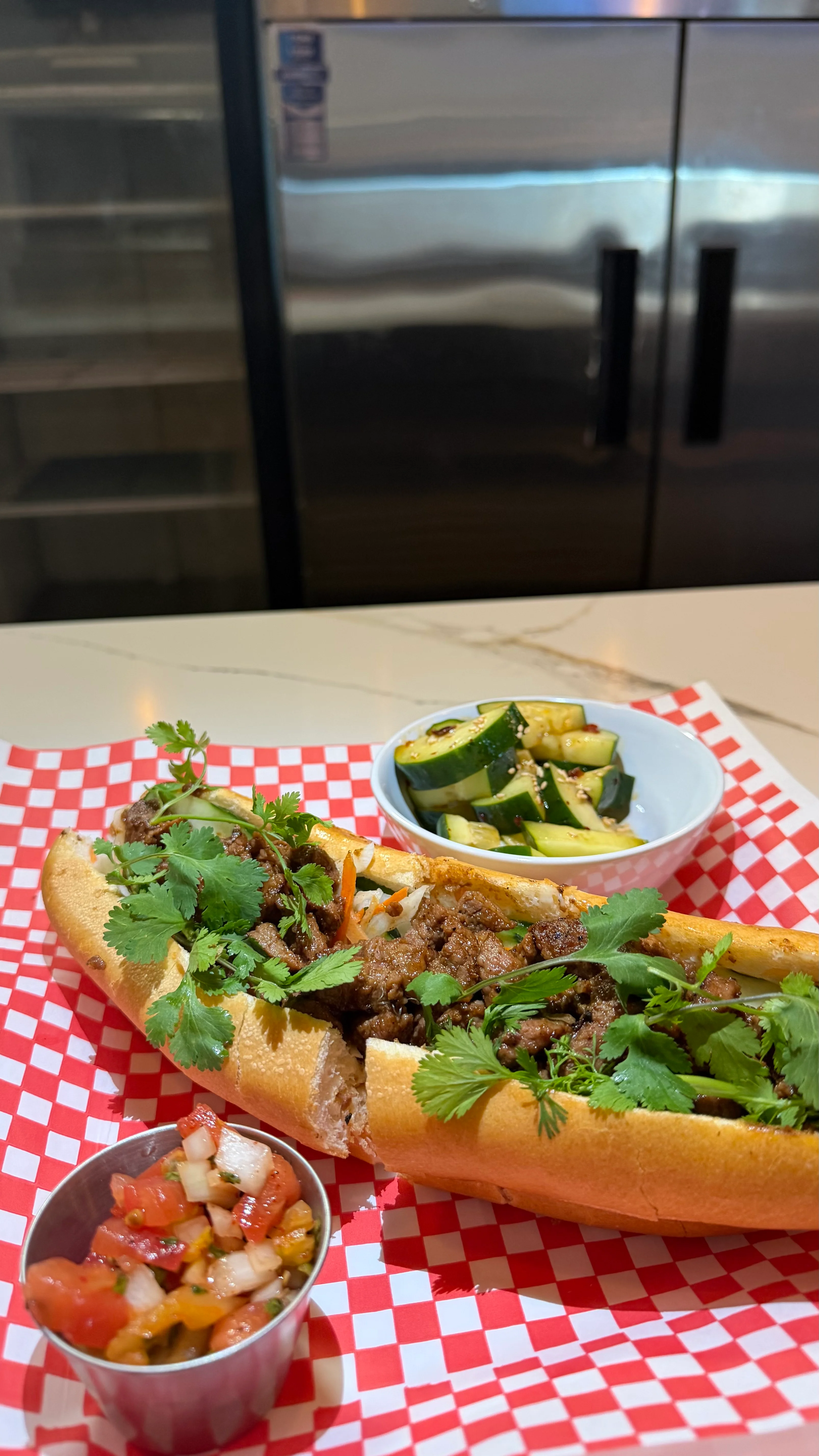 A close-up of a hot dog filled with meat and topped with cilantro, served on a checkered paper with a small bowl of cucumber salad and salsa.