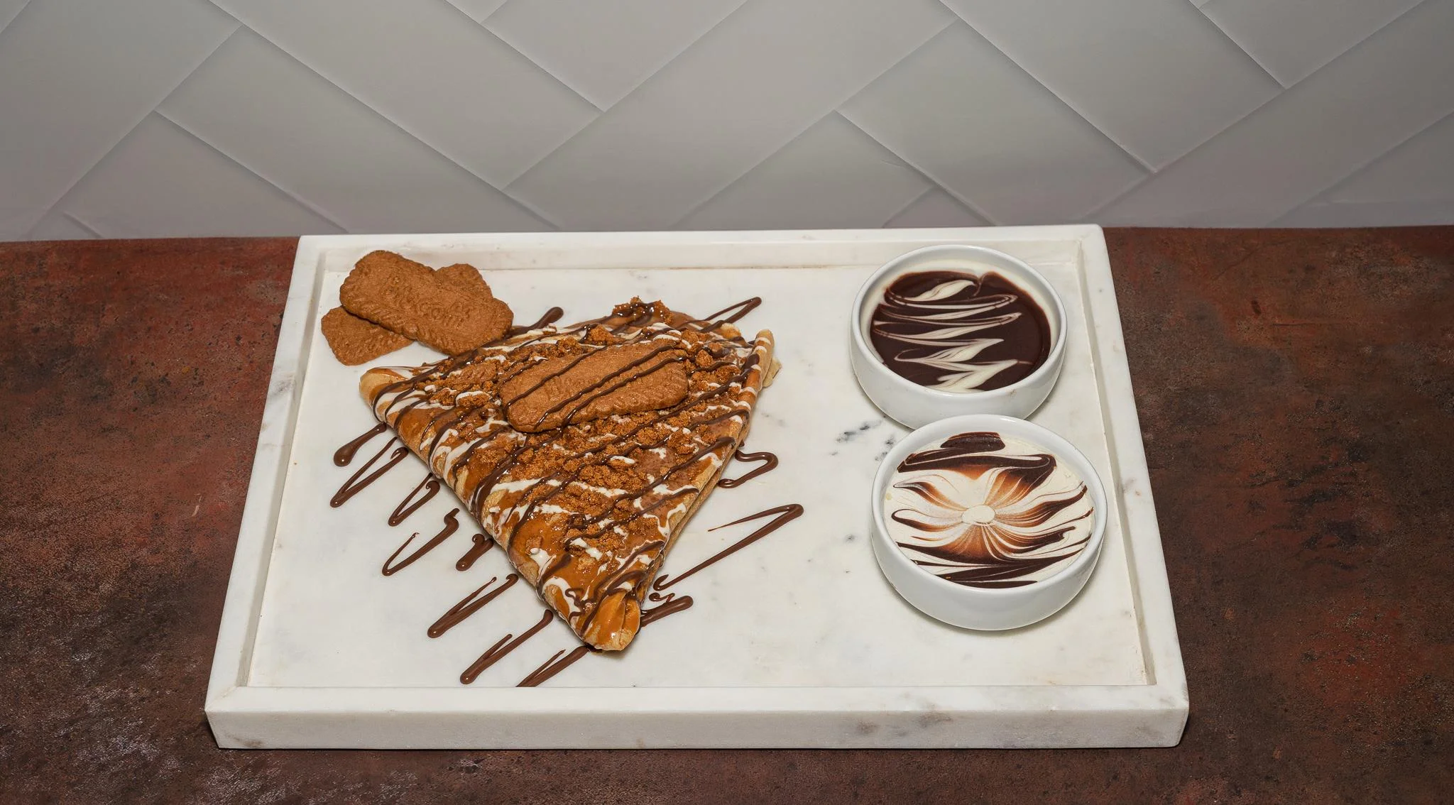 A delicious dessert crepe topped with caramel sauce, crushed cookies, and drizzled chocolate, served with two bowls of chocolate sauce.