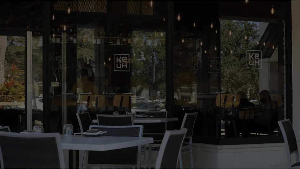The exterior patio of Ke-uH Boca Raton featuring outdoor dining tables and chairs set up in front of large, dark-tinted glass windows showing indoor seating and hanging pendant lights.