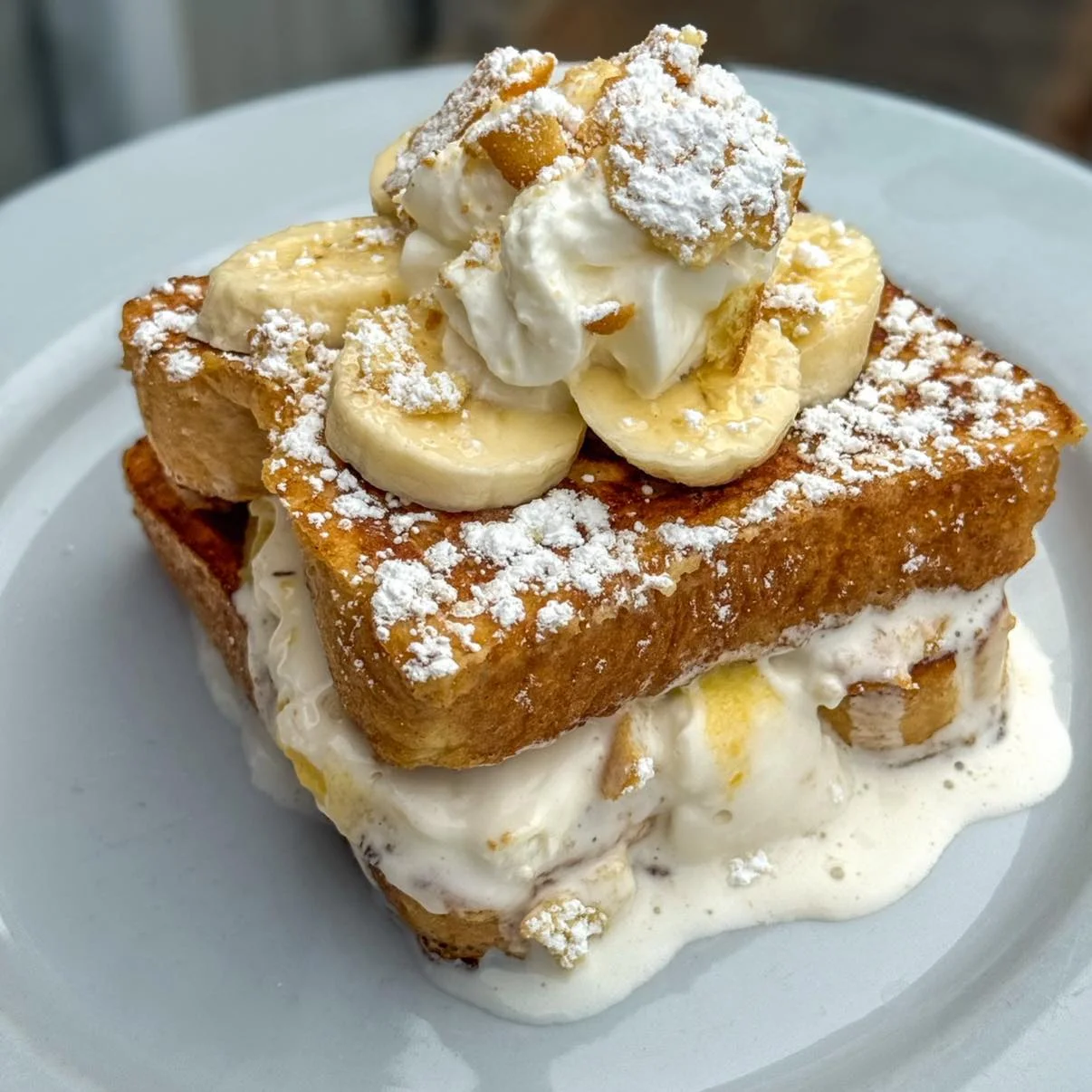 A decadent dessert featuring layers of French toast, cream, and banana slices, dusted with powdered sugar.