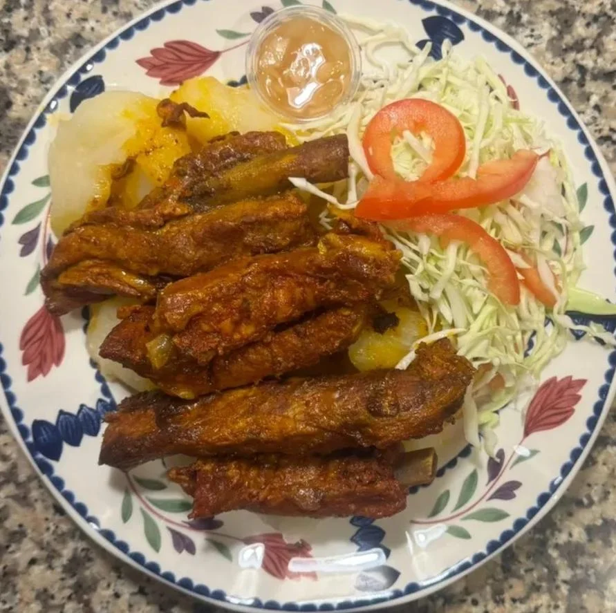 A plate featuring grilled meat ribs served with yucca, salad, and a small container of sauce, surrounded by fresh vegetables.