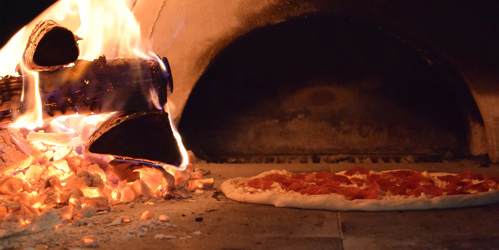 Pepperoni pizza in a wood-fire oven with logs and a flame on the left side
