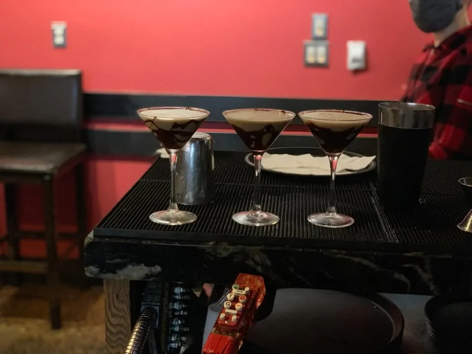 Three chocolate martinis elegantly presented in a row, served on a bar counter with a vibrant red background. Each glass has a chocolate drizzle design.