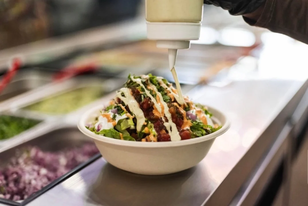 A bowl filled with fresh ingredients, topped with sauce, being prepared in a food service setting.