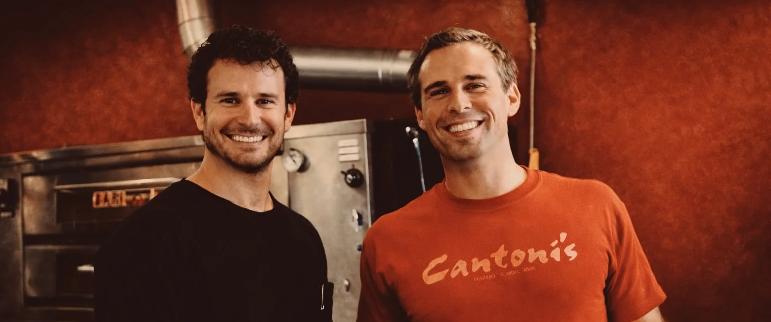 Two smiling individuals standing in a kitchen environment with a metal oven in the background.