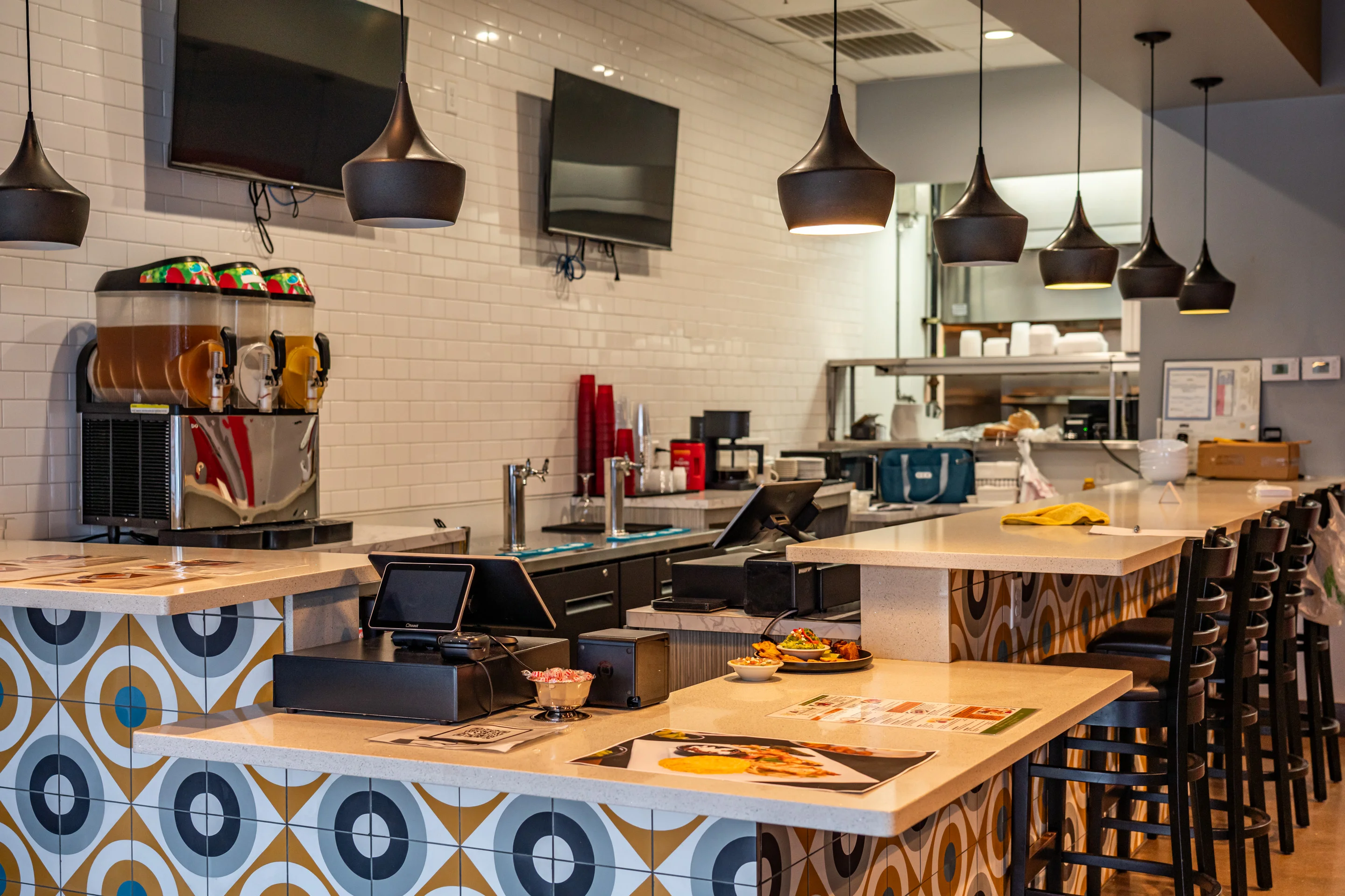 A modern restaurant interior with a sleek counter, beverage dispensers, and hanging pendant lighting.