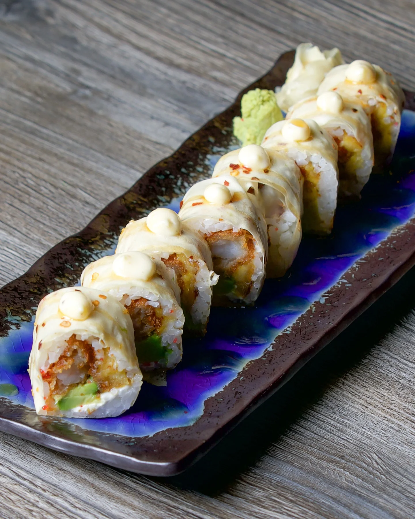 A long row of specialty sushi rolls filled with crunchy tempura, avocado, and cream cheese, wrapped in a light soy paper instead of seaweed, and topped with a dot of creamy mayo on each piece, served on a dark glazed ceramic plate with purple accents alongside wasabi and ginger.