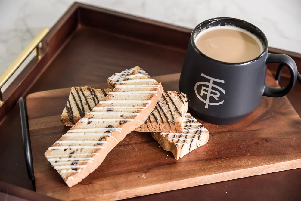 A black mug filled with coffee sits on a wooden tray beside four biscotti drizzled with white and dark chocolate. The scene conveys a cozy, inviting breakfast.