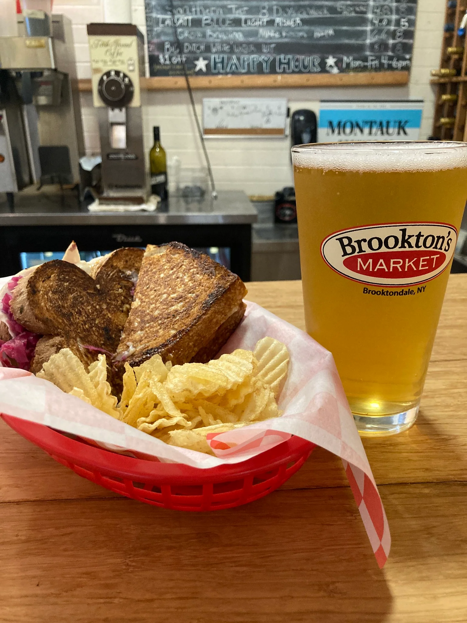 A toasted sandwich served with potato chips in a red basket, alongside a pint of beer with a logo on the glass. The background features a chalkboard menu.