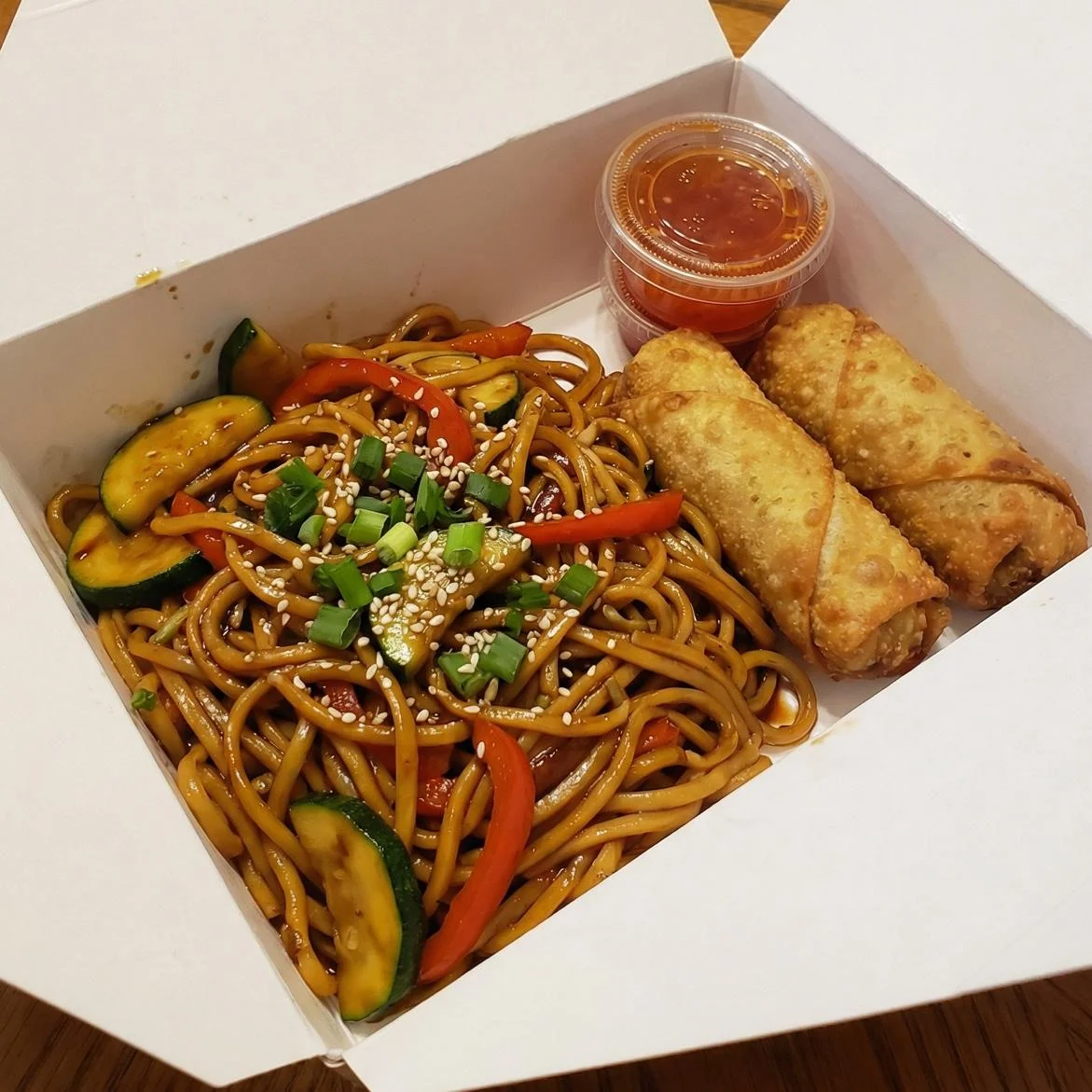 A takeout box containing stir-fried noodles with vegetables, two spring rolls, and a small container of dipping sauce.