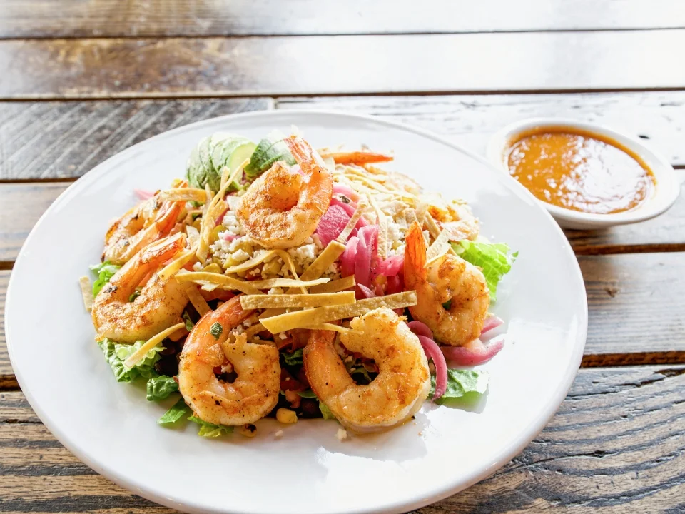 A vibrant shrimp salad featuring grilled shrimp on a bed of lettuce, garnished with crispy tortilla strips and served with a tangy dipping sauce on the side.