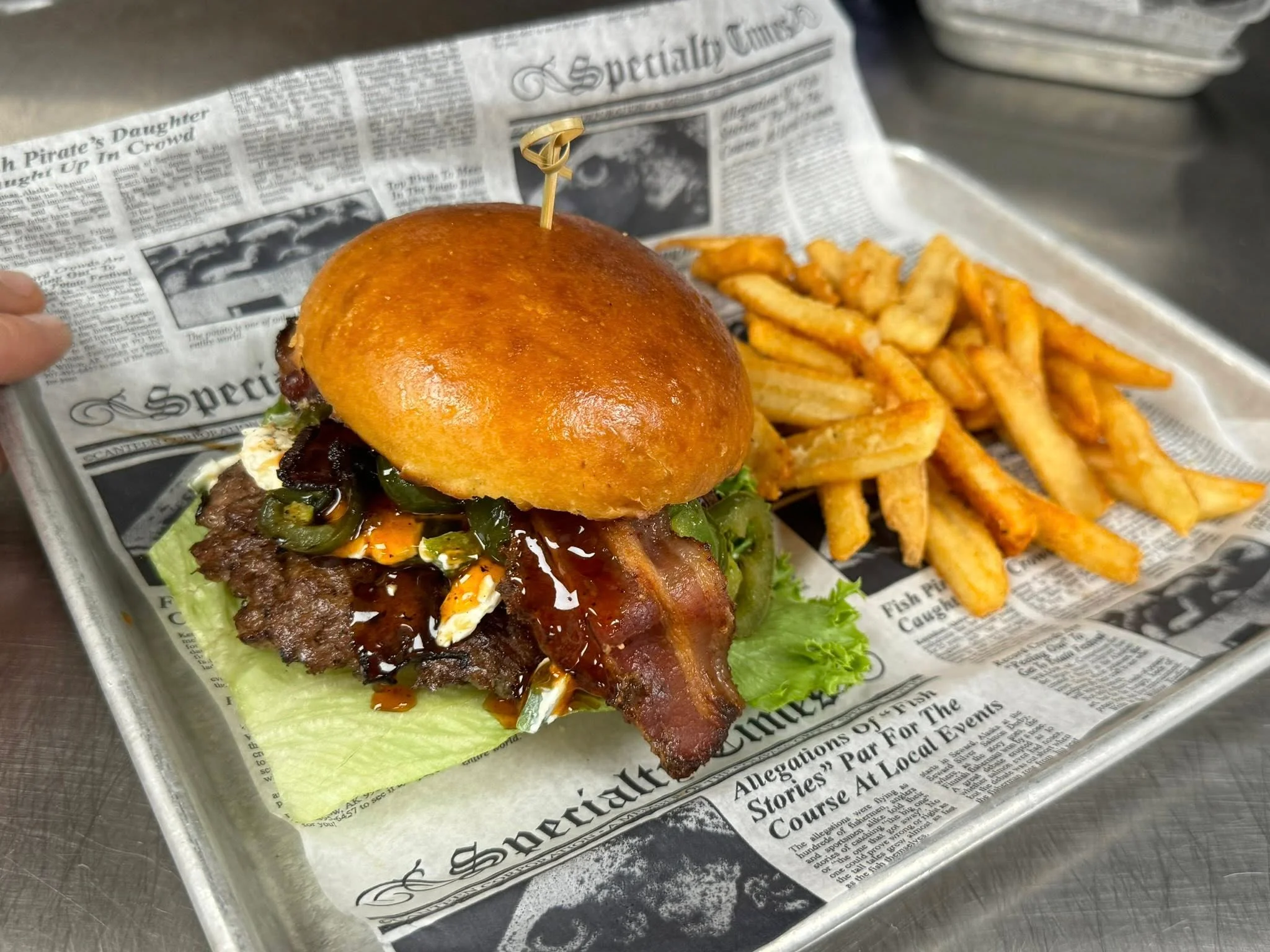 A close-up of a bacon cheeseburger with lettuce, onion, and melted cheese on a brioche bun, served with seasoned fries.