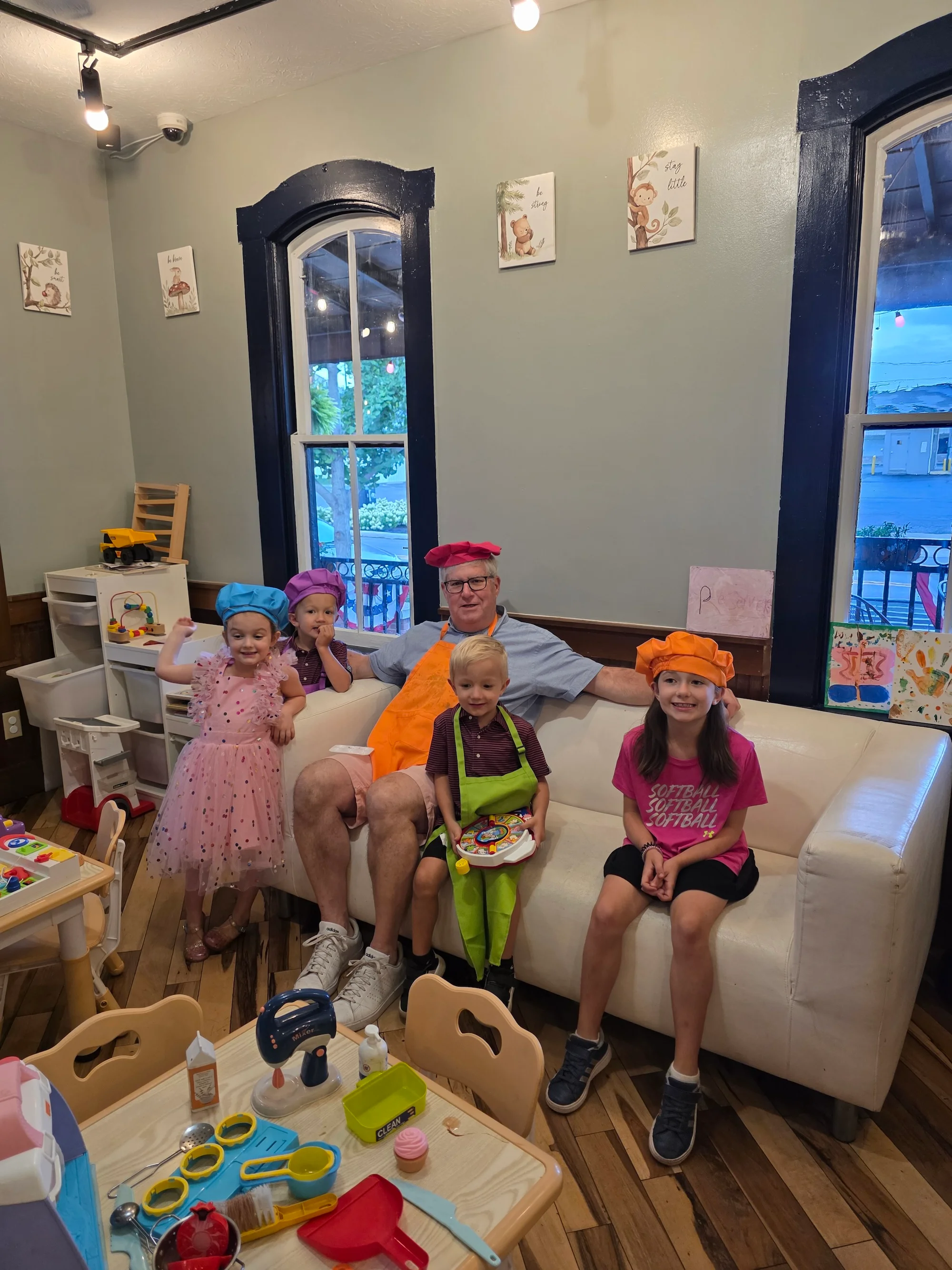 A group of children and an adult, all wearing colorful chef hats, are engaged in playful activities in a cozy indoor space filled with toys and decorations.