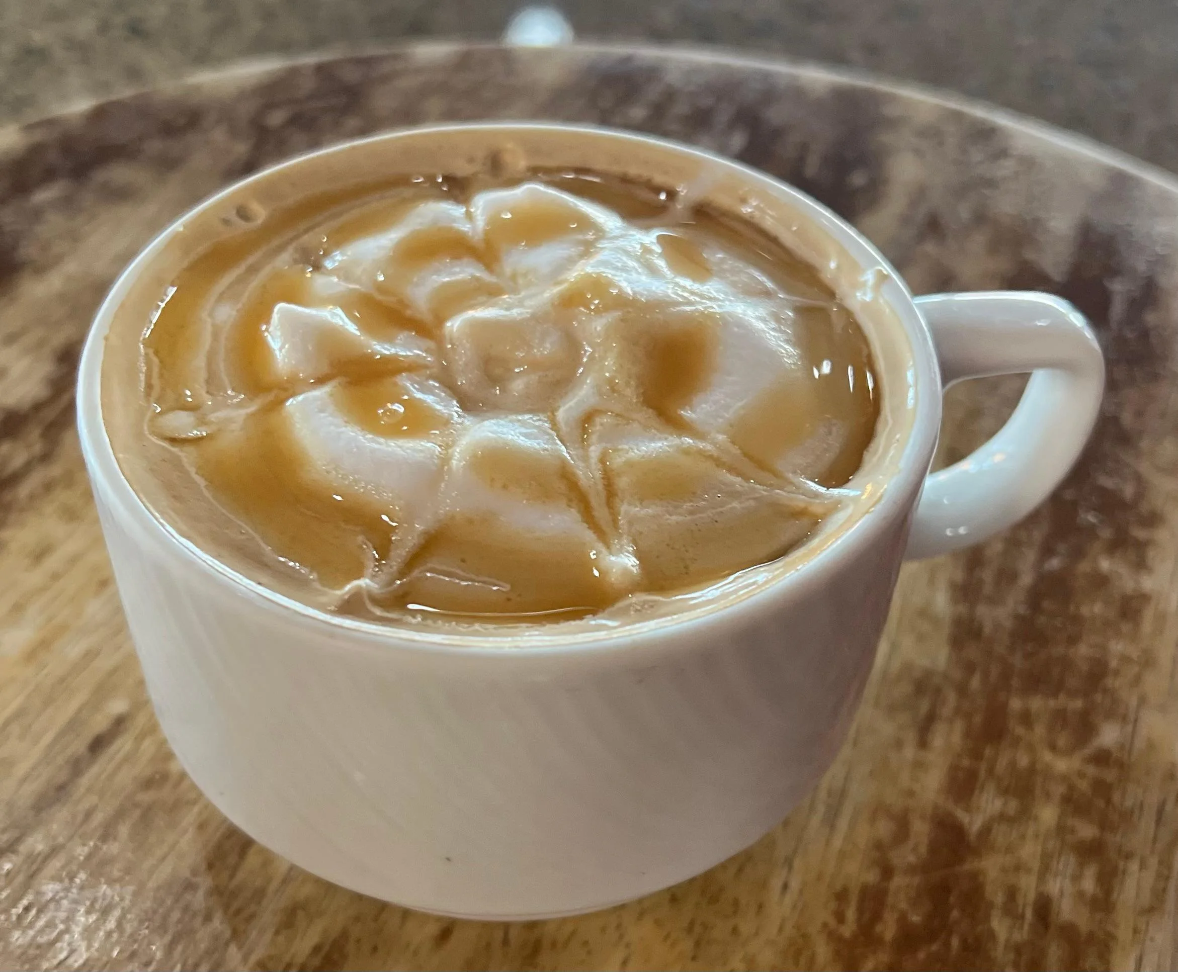 A small white cup filled with a caramel-colored coffee drink topped with a decorative foam pattern.