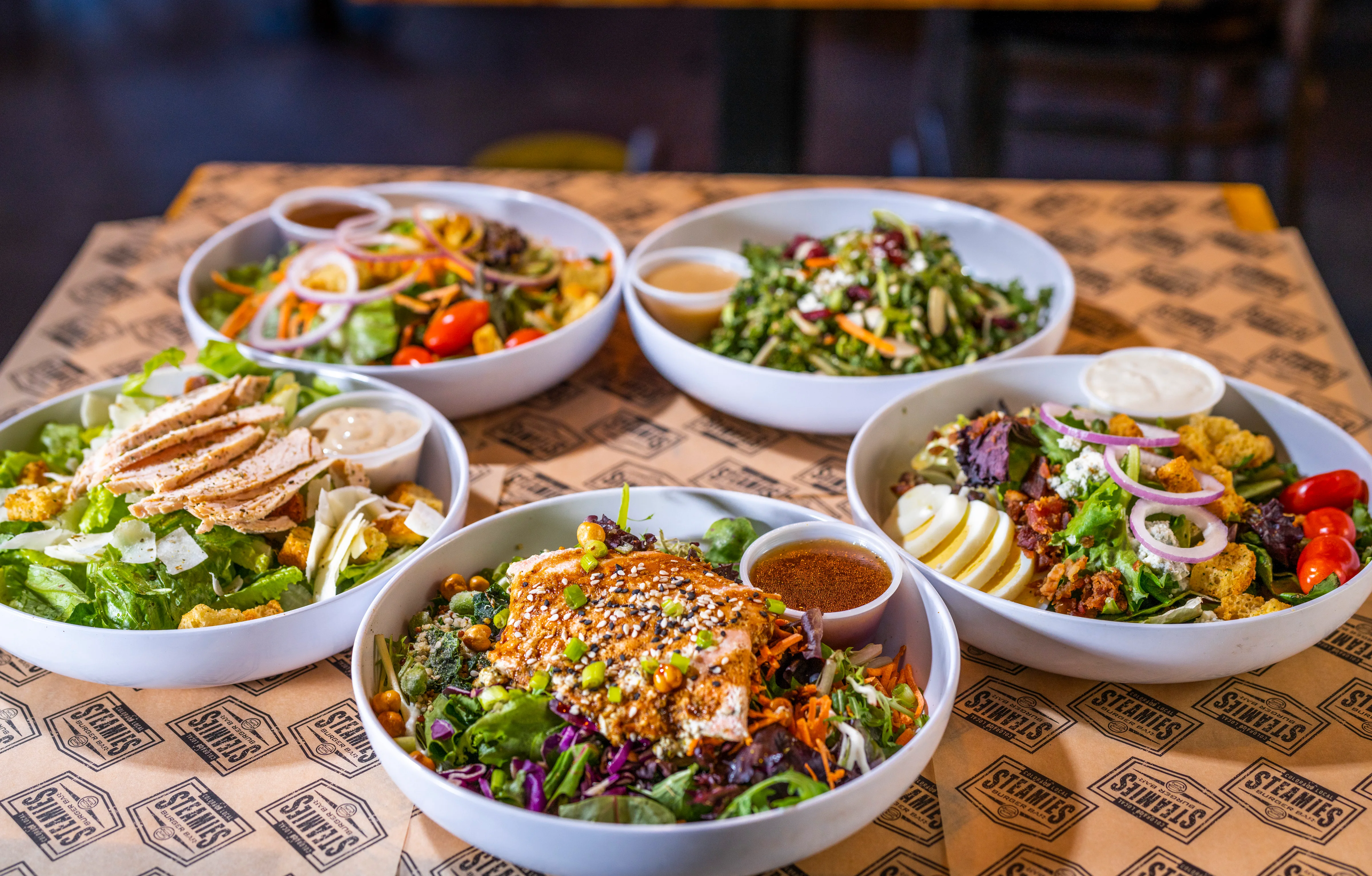 A variety of fresh salads served in large bowls, showcasing colorful vegetables and toppings, garnished on a branded table.