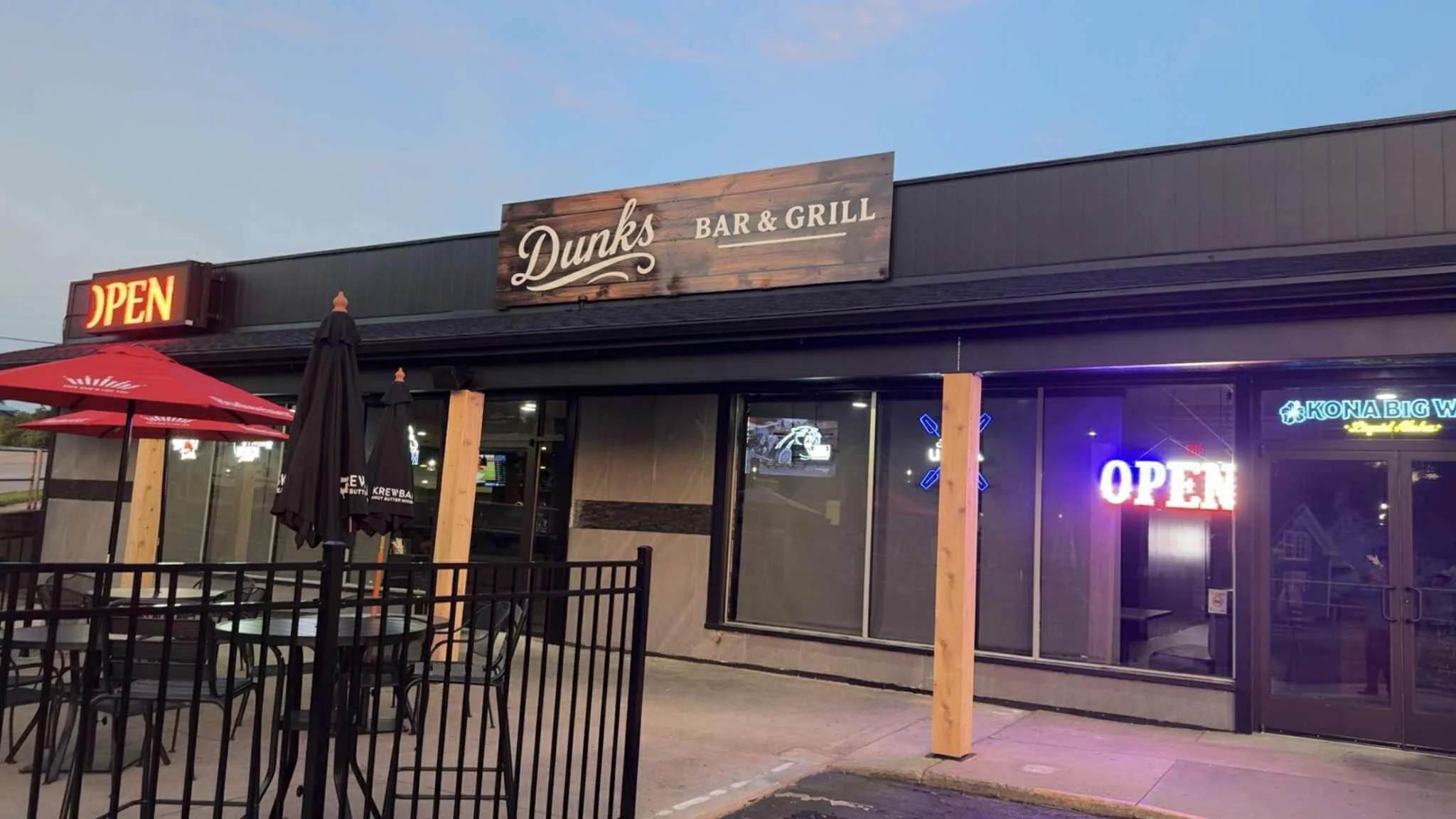 Exterior view of Dunks Bar & Grill with patio seating, umbrellas, neon “Open” signs, and a wooden sign above the entrance.