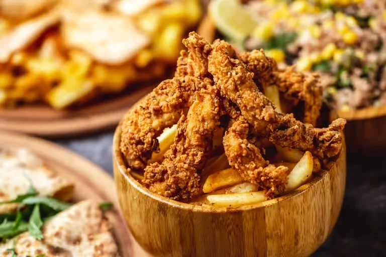 A bowl filled with crispy fried chicken strips and fries, surrounded by various other food items on wooden plates.