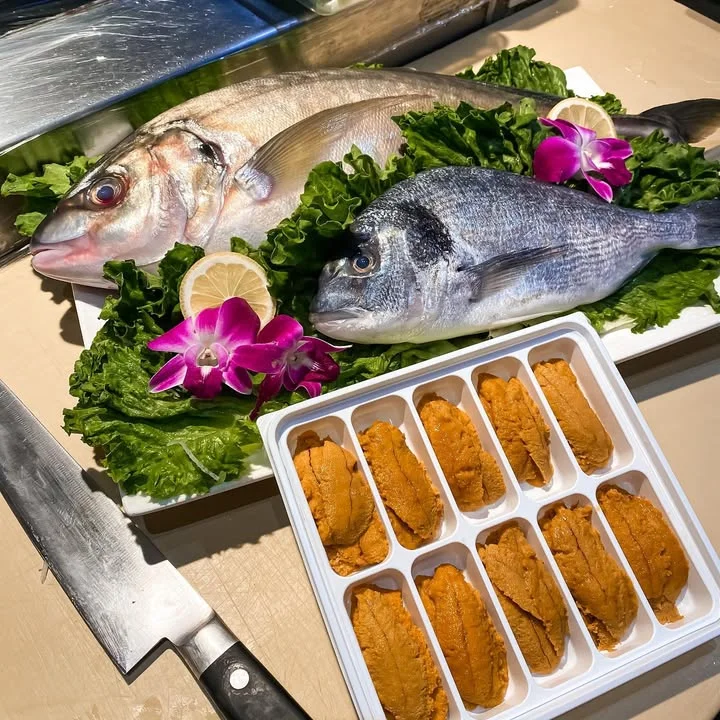 A display of fresh fish, including two whole fish and a tray of sea urchin, arranged on a bed of lettuce with a lemon and flowers for decoration.