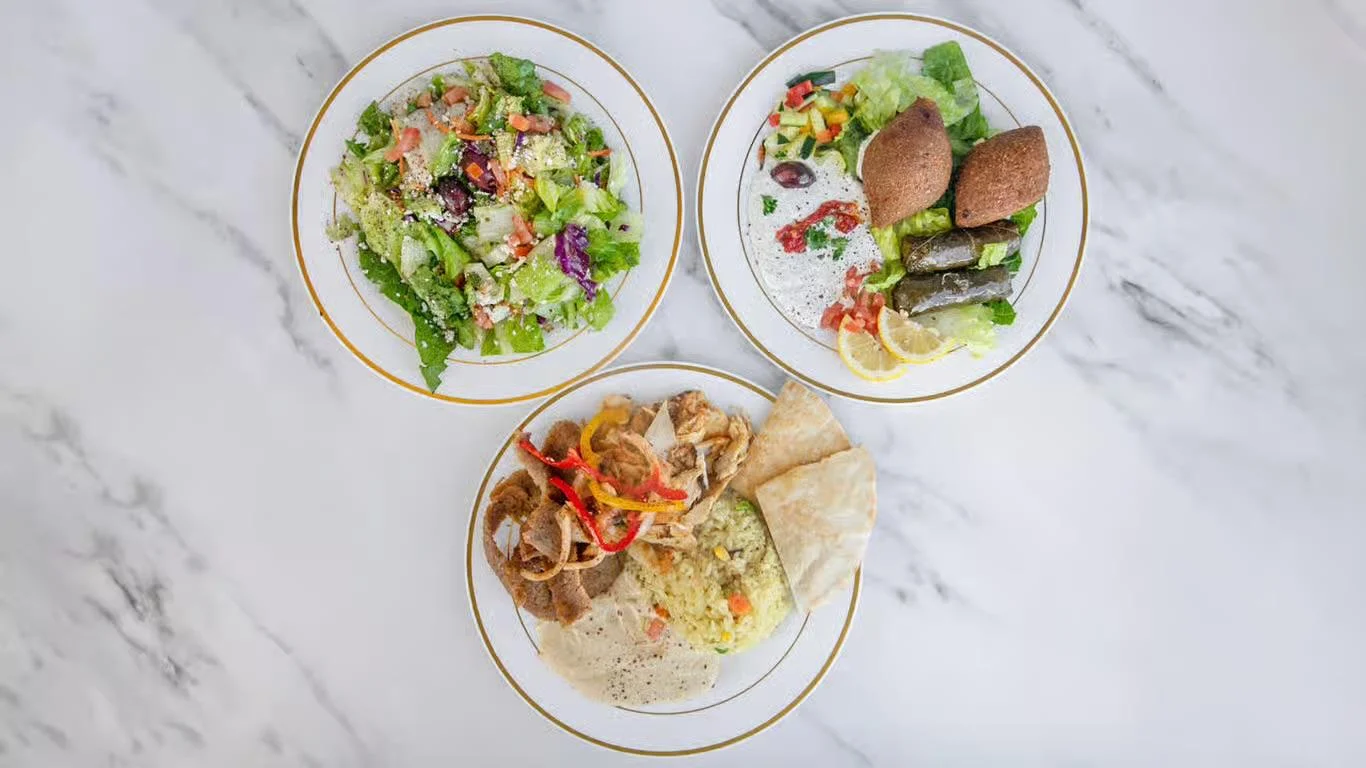 An assortment of Mediterranean dishes including salads and meat served on white plates with gold trim, arranged neatly on a marble table.
