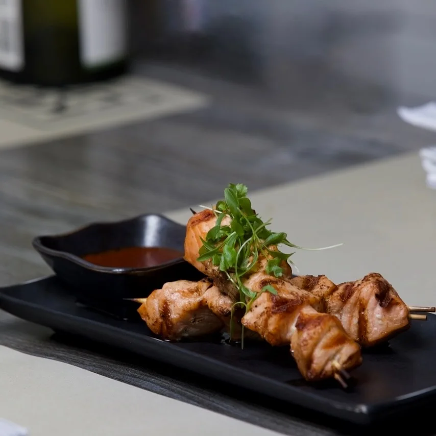 Two grilled salmon skewers with distinct grill marks, garnished with fresh microgreens and resting on a long black rectangular plate alongside a small dark bowl of dipping sauce on a restaurant table.
