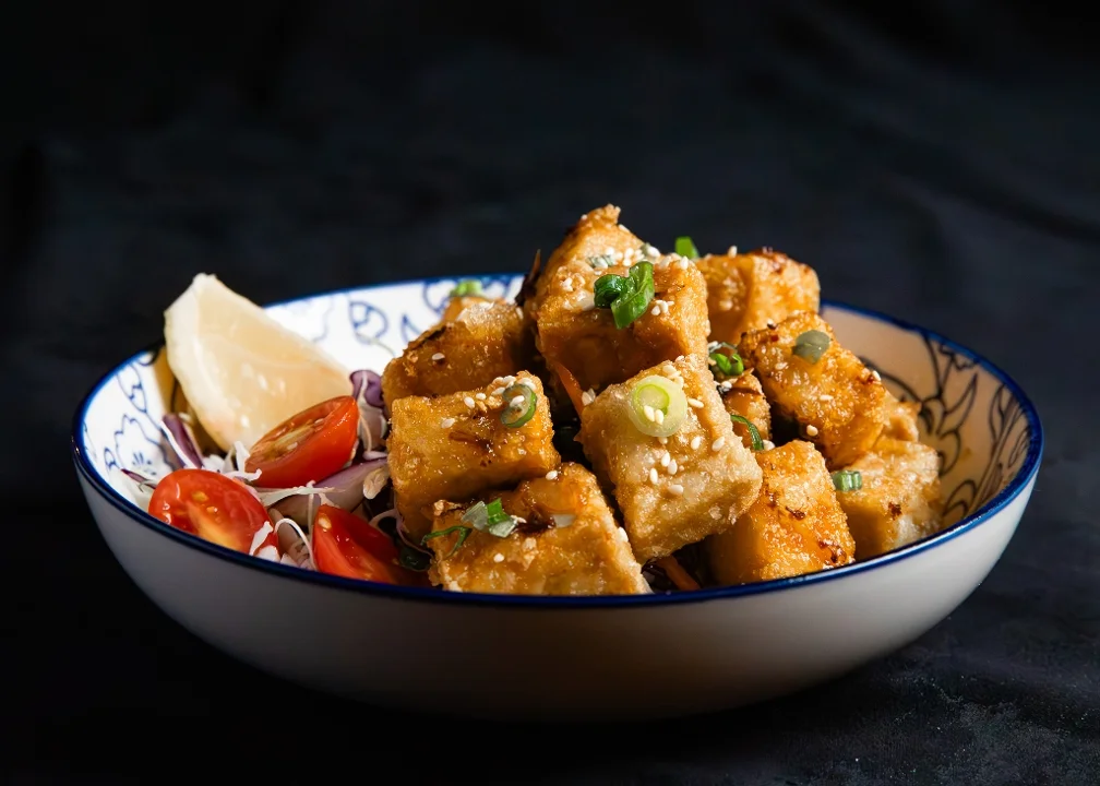 A plate of crispy fried tofu cubes, garnished with green onions, surrounded by tomatoes and a lemon slice.