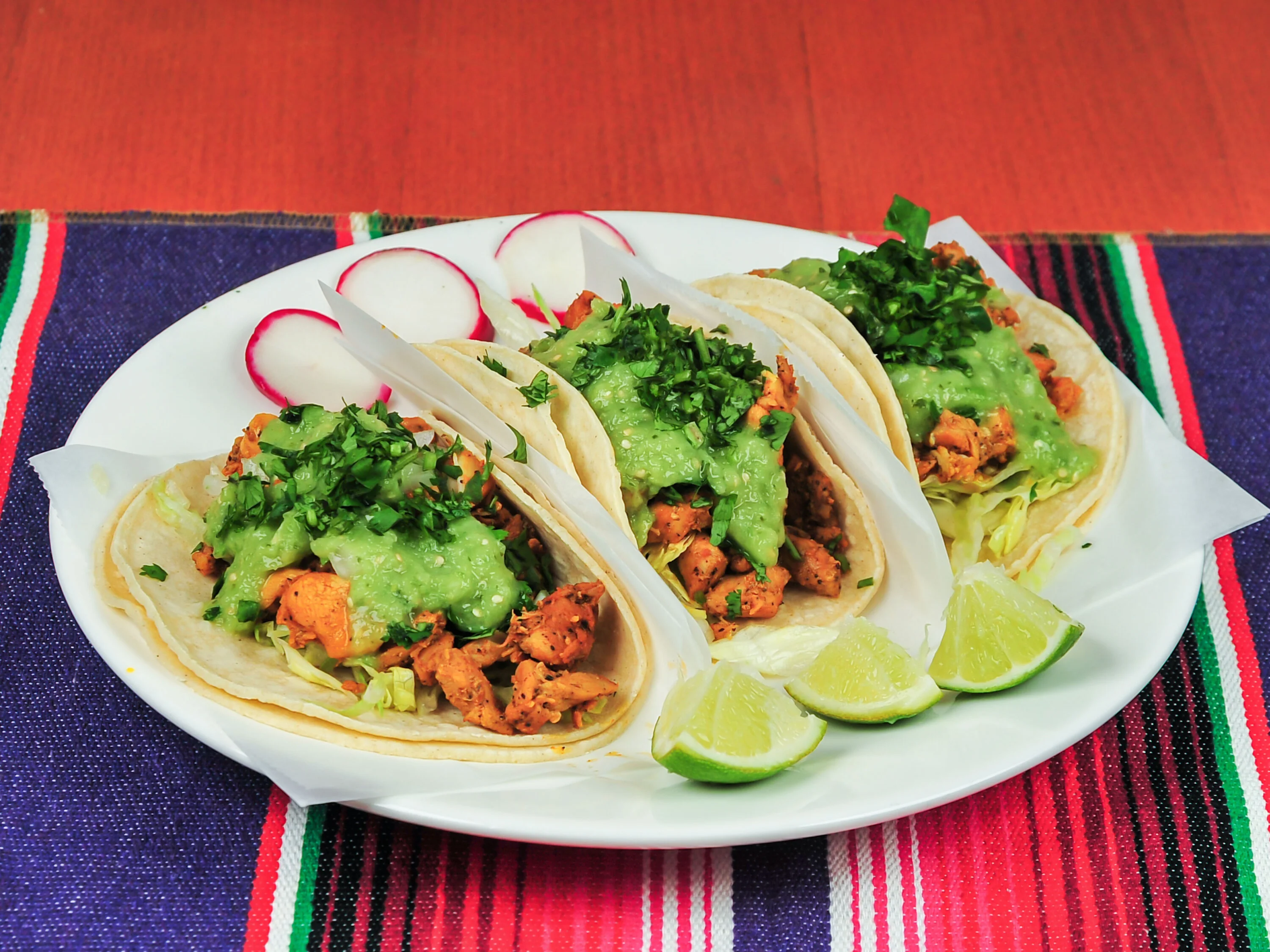 Three tacos filled with seasoned meat, topped with green sauce and chopped cilantro, accompanied by lime wedges and radishes.