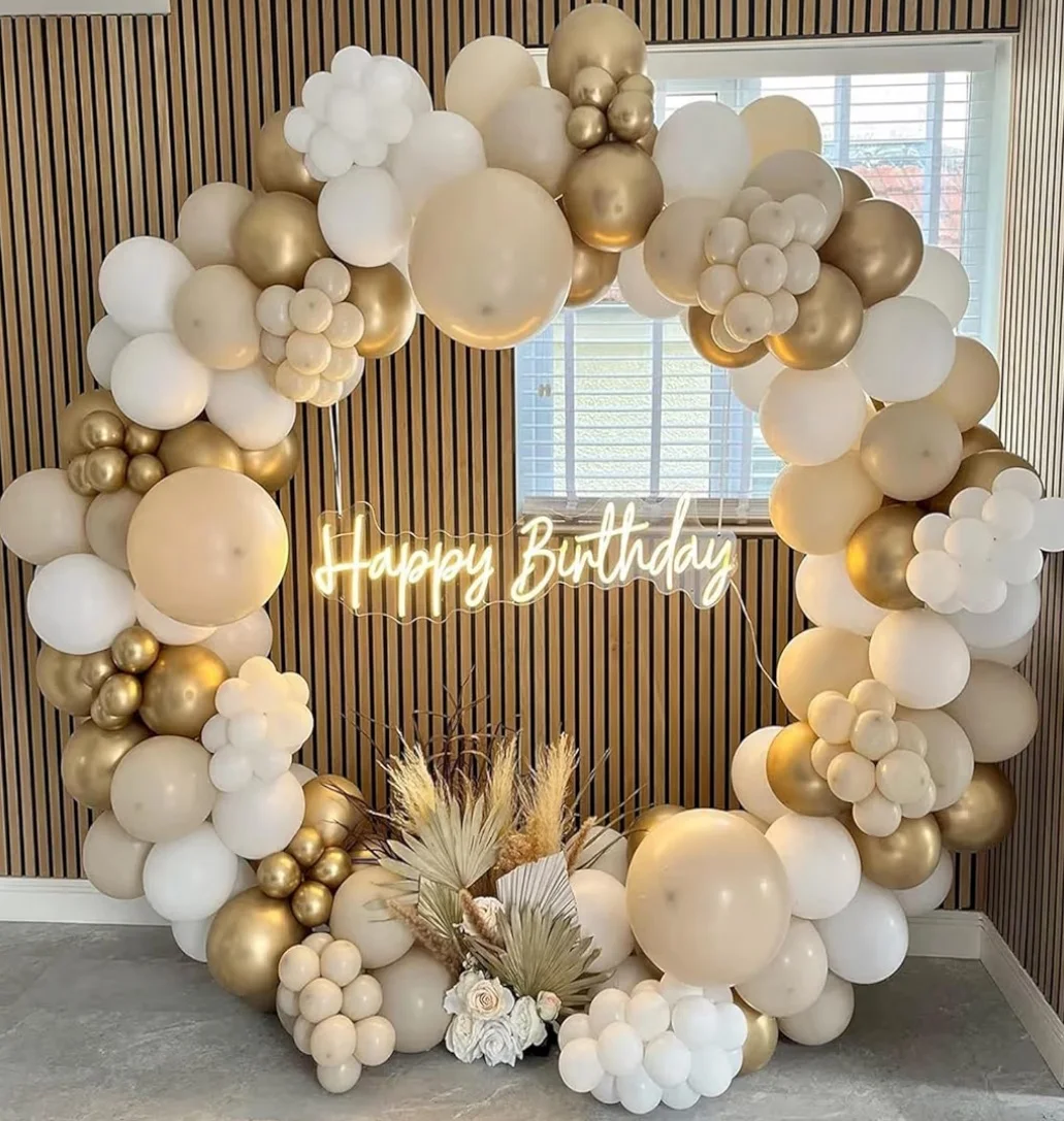 A decorative arch made of balloons in shades of gold and white, featuring a glowing 'Happy Birthday' sign and an arrangement of dried flowers at the base.