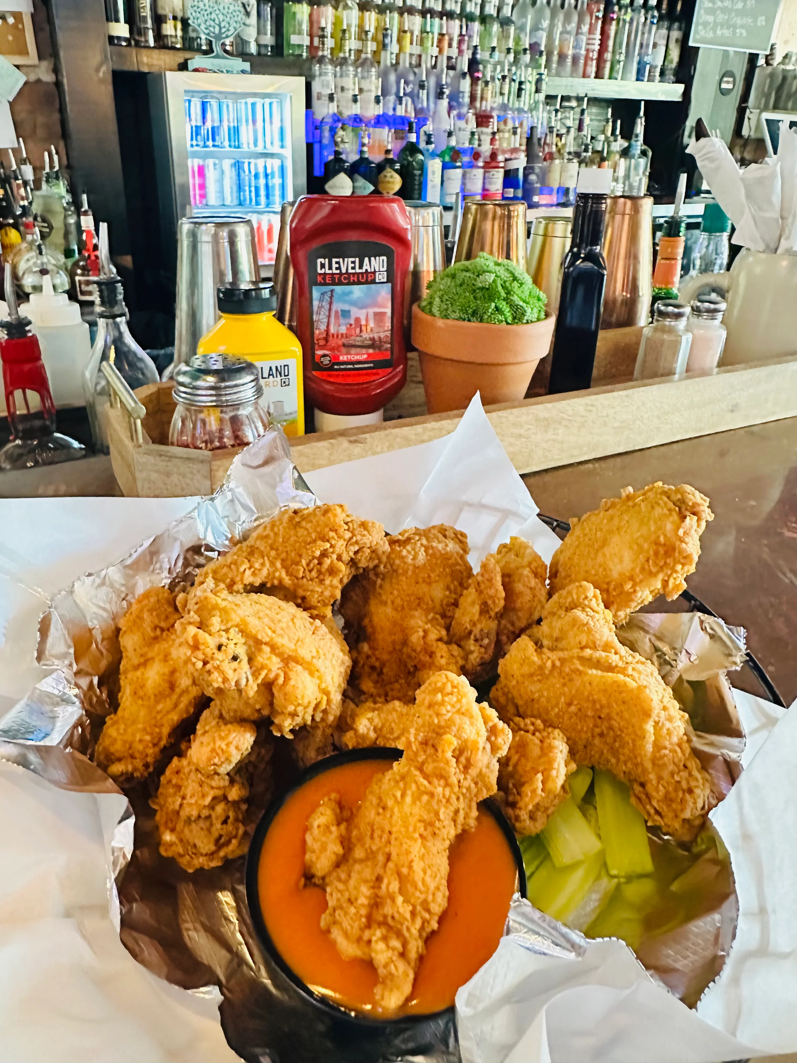 A platter of crispy fried chicken wings served with a side of orange dipping sauce and celery sticks. The backdrop includes a well-stocked bar.