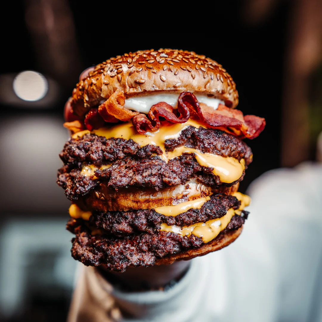 A towering triple patty burger topped with melted cheese and crispy bacon, showcased in hand.