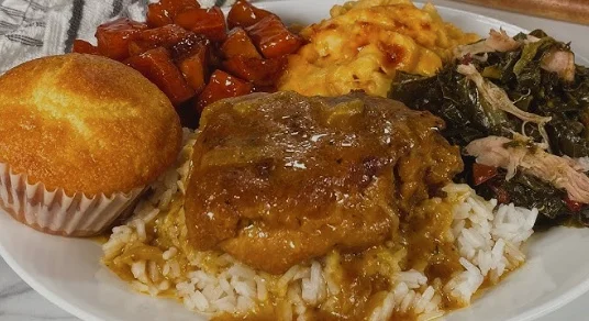 A plate of soul food with rice and gravy, collard greens with smoked turkey, candied yams, baked macaroni and cheese, and cornbread.