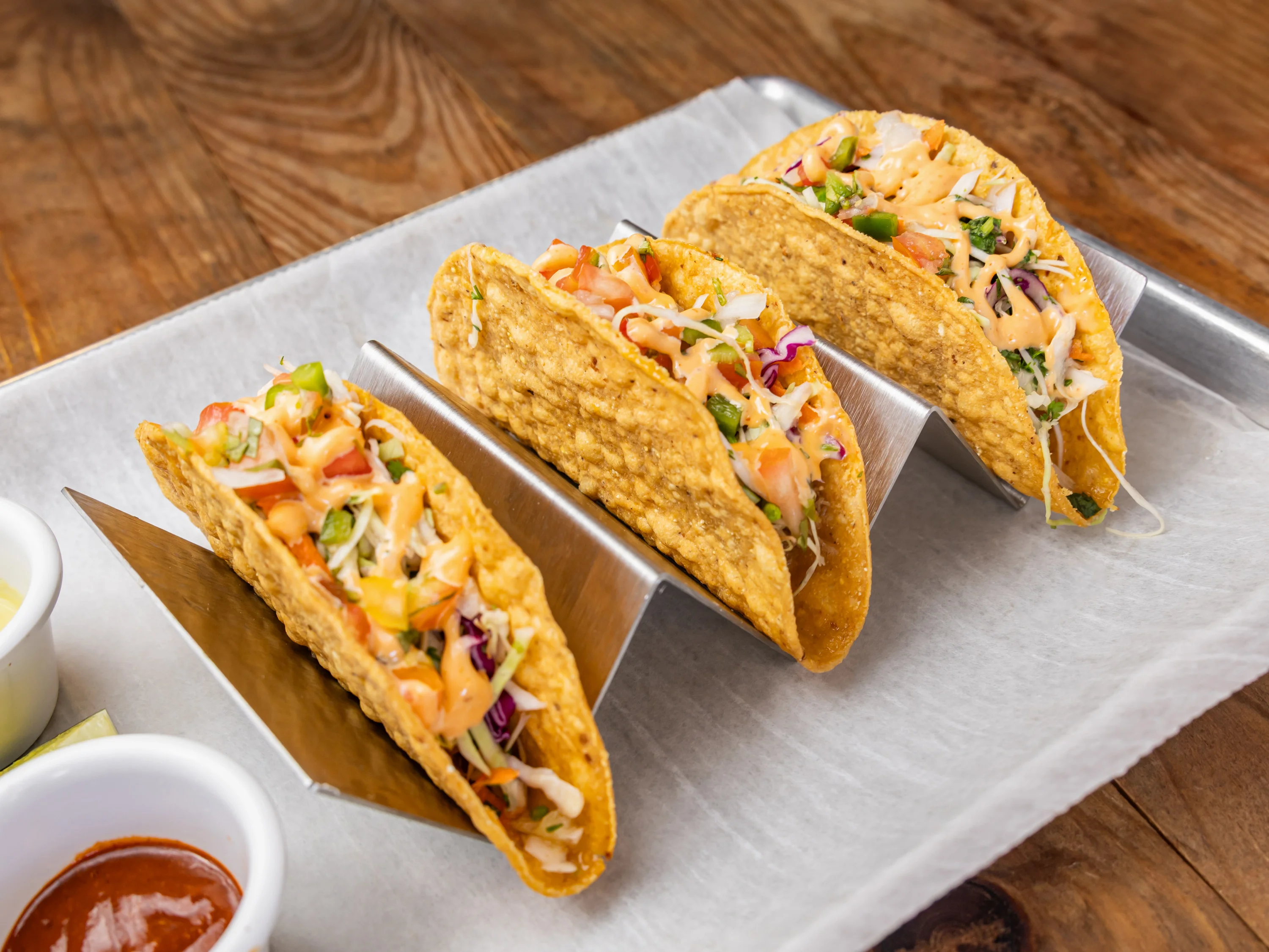 Three tacos filled with a mix of vegetables and sauce, served in a taco holder. Accompanied by small bowls of salsa.