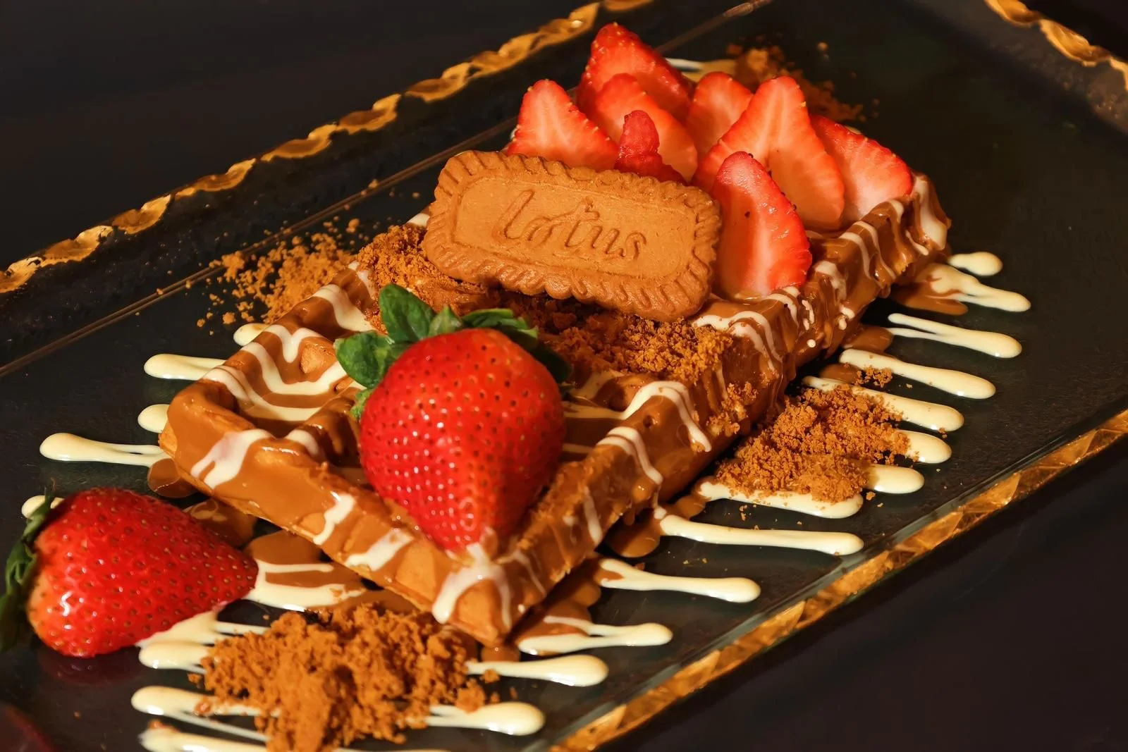 A decadent dessert featuring layers of chocolate, strawberries, and a biscuit on top, garnished with drizzles of white sauce and sprinkled crumbs.
