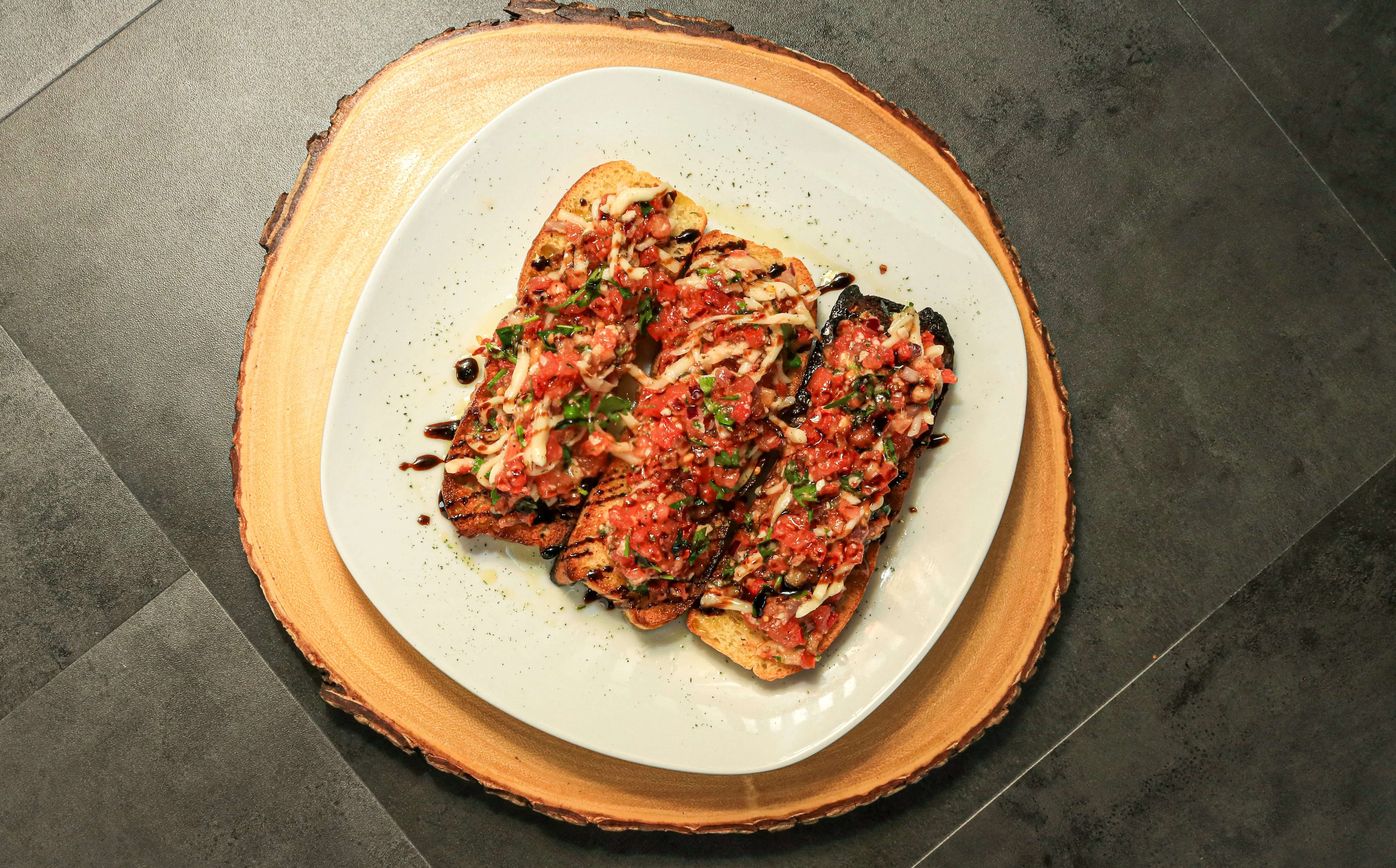 Grilled bread slices topped with a mixture of diced tomatoes, herbs, and drizzled with a balsamic reduction. The dish is presented on a wooden serving platter.