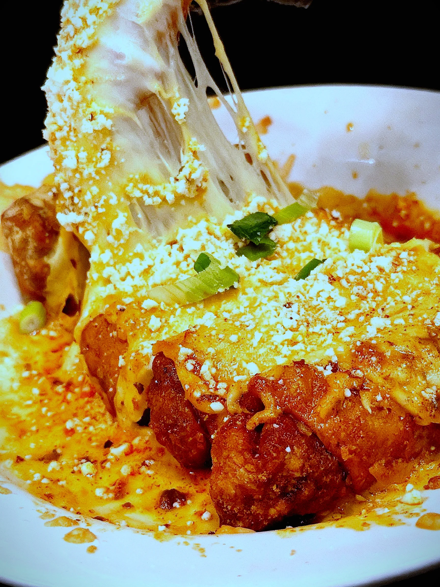 A close-up of a cheesy dish garnished with scallions and crumbled cheese, showcasing a stretchy cheese pull.