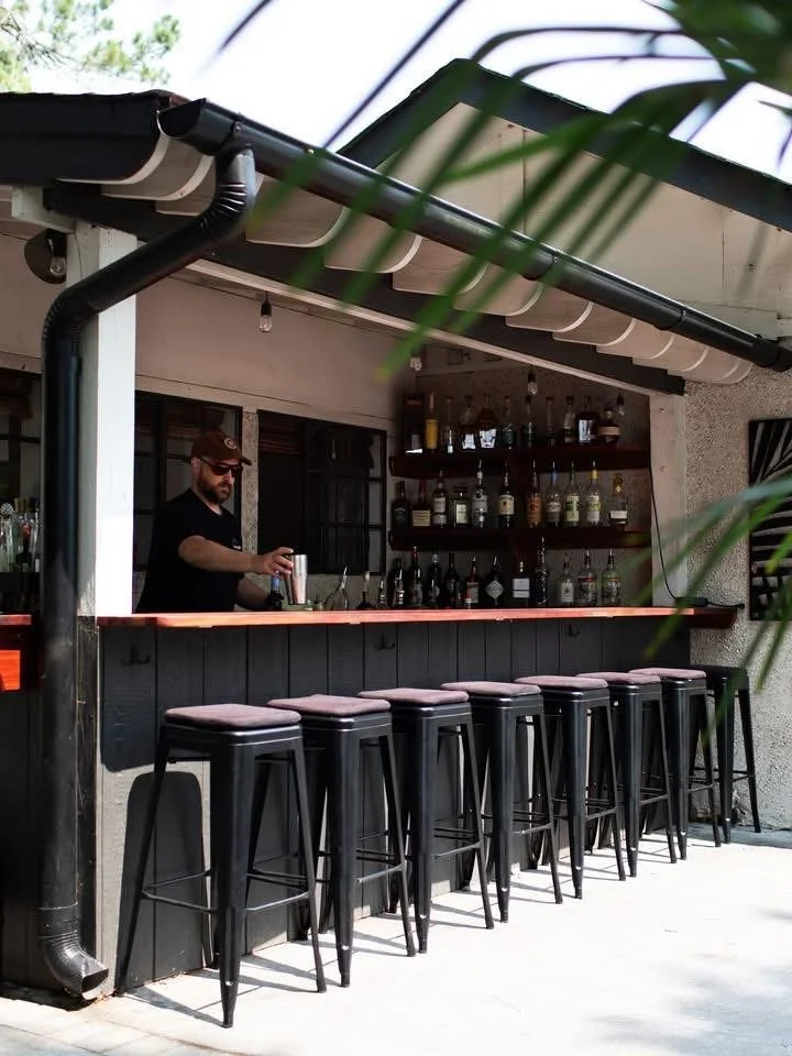 An outdoor bar with a bartender mixing drinks, featuring high stools and a shelf of various bottles.
