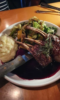 A serving of grilled rack of lamb alongside mashed potatoes and a side of mixed vegetables on a white plate.